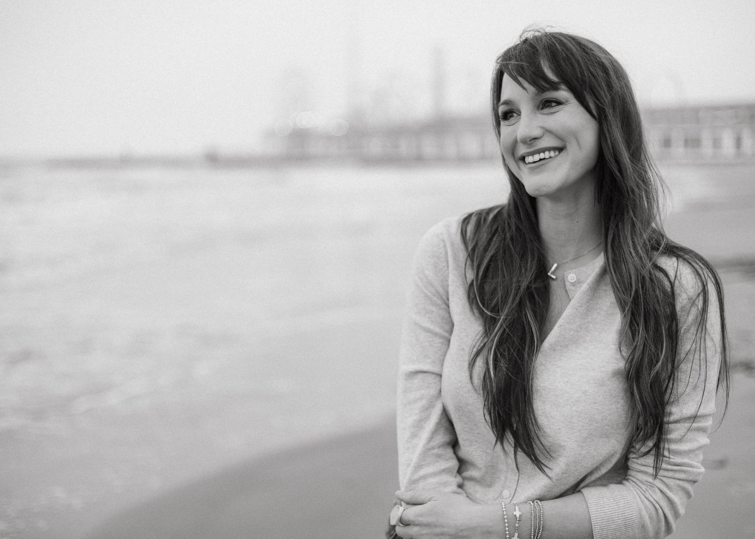 Smiling young woman with long dark hair posing on a beach in a black and white portrait photo.