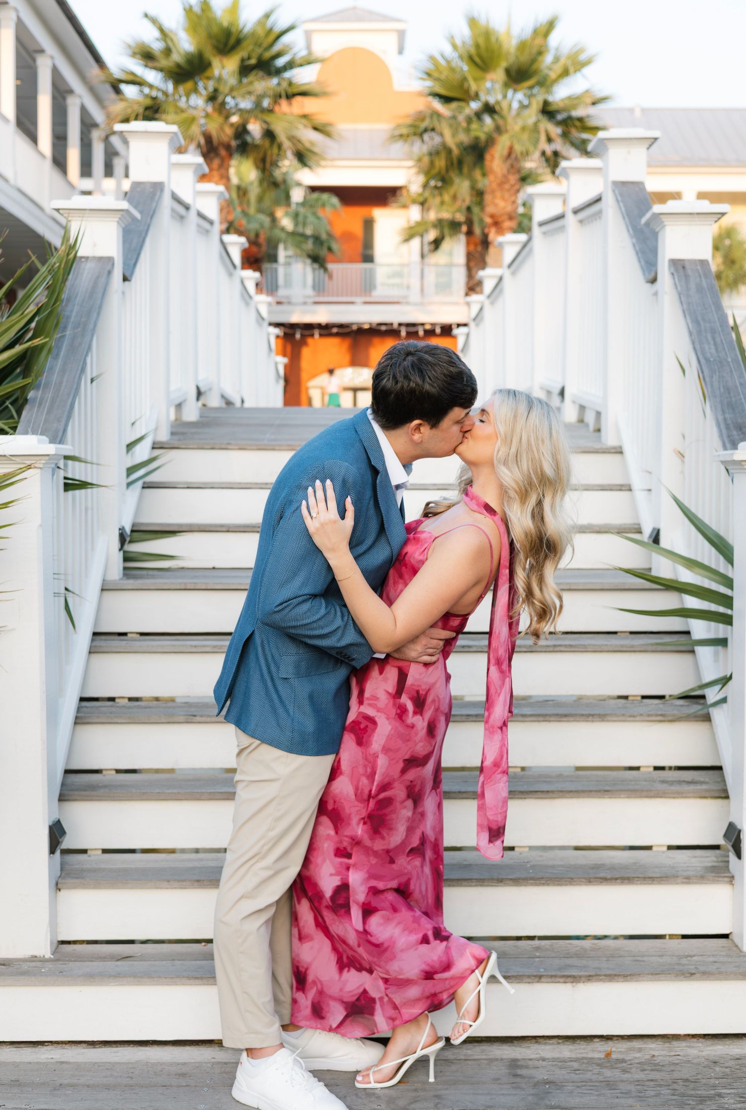 Couple sharing a romantic kiss in front of white stairs, man in blue blazer, woman in pink floral dress.