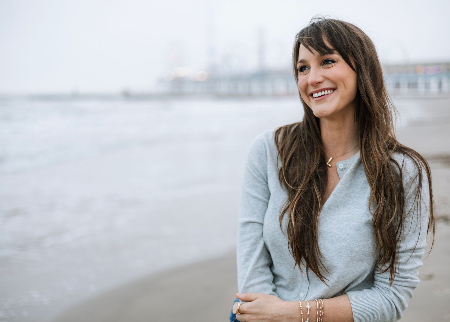 Smiling young woman with long brown hair on a beach, wearing a gray sweater with a city pier in the background.
