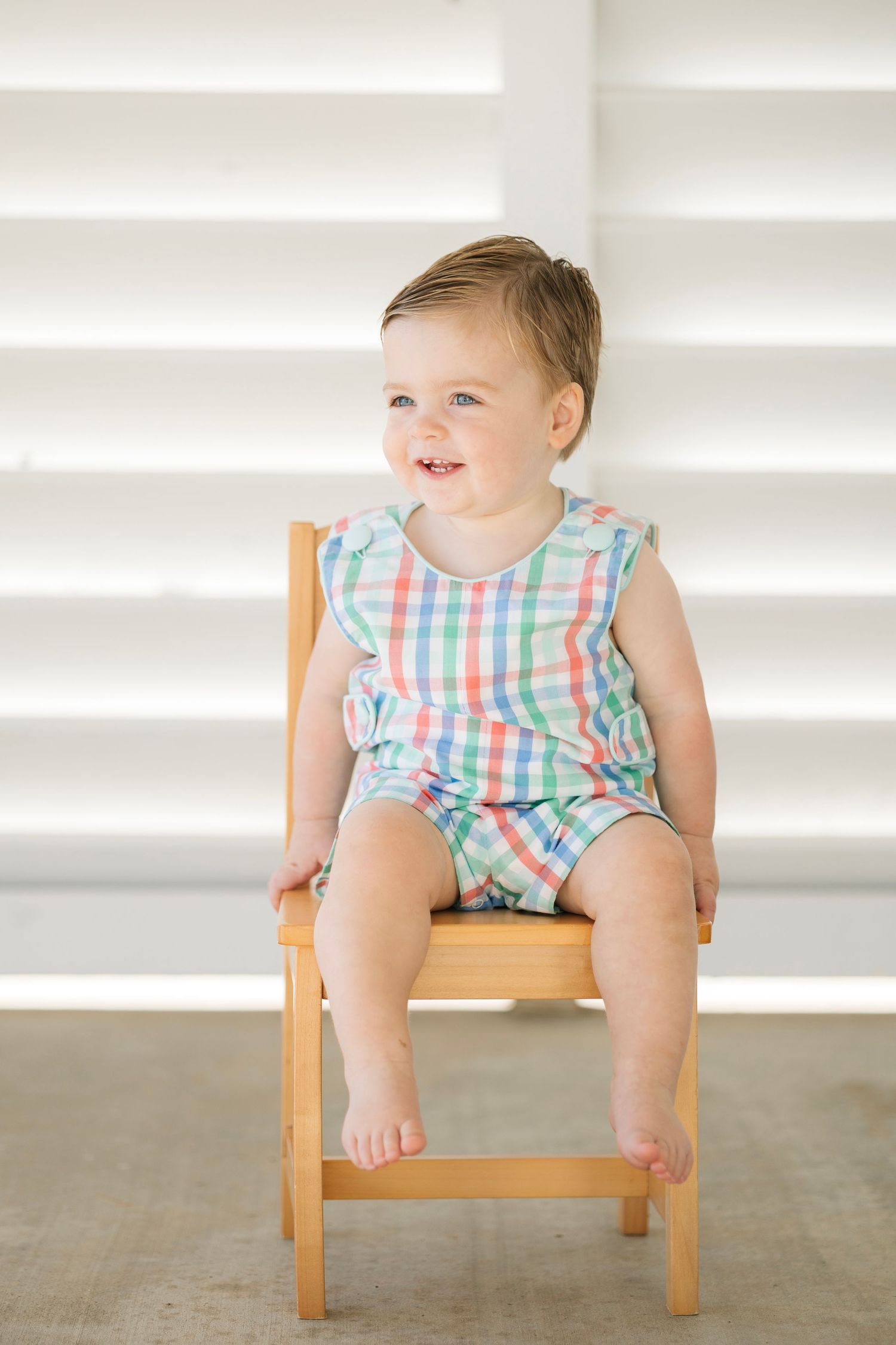 A happy baby in a colorful plaid romper sits smiling on a small wooden chair against a white background.