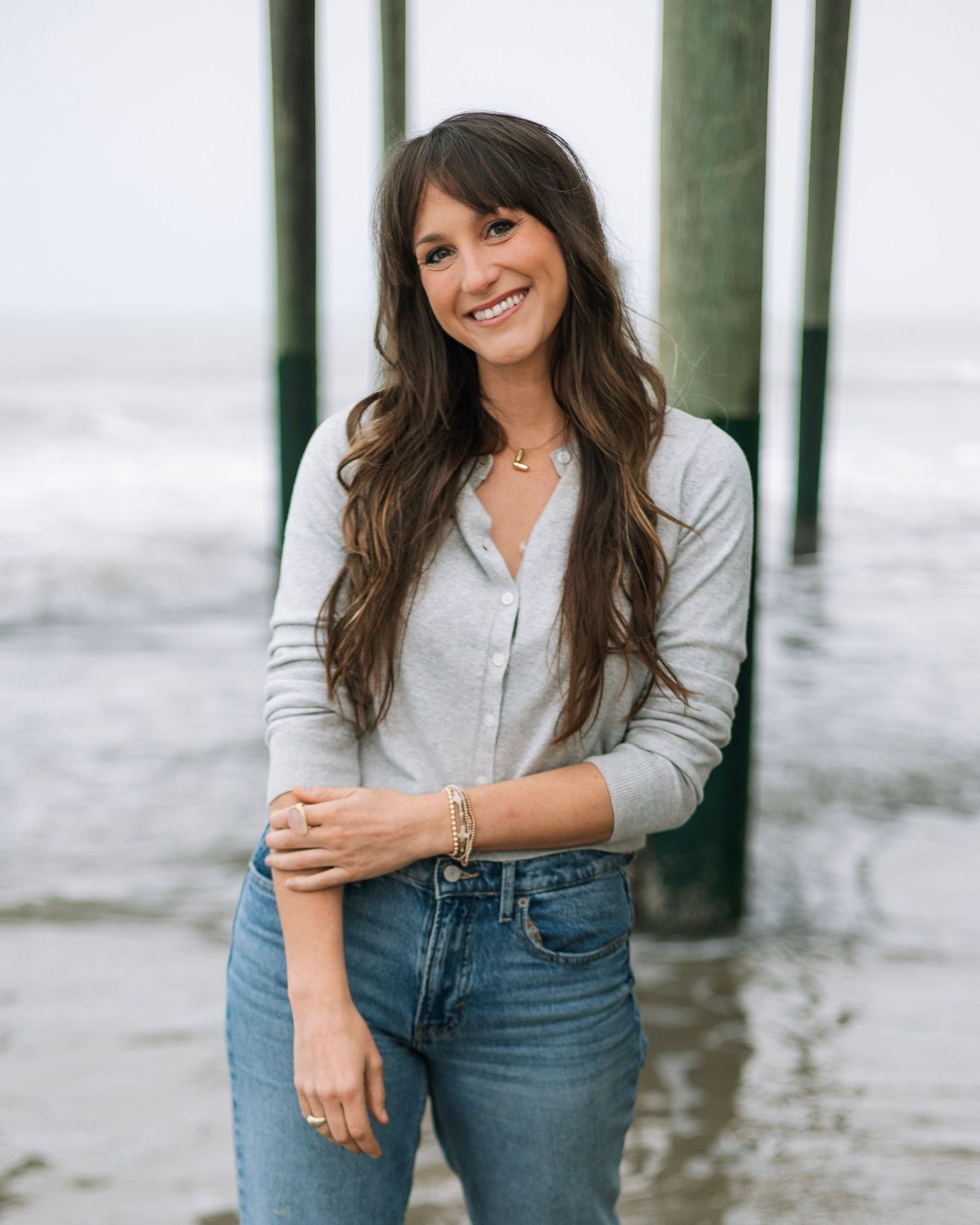 Smiling brunette woman in gray cardigan and jeans posing at beach pier with ocean waves in background.