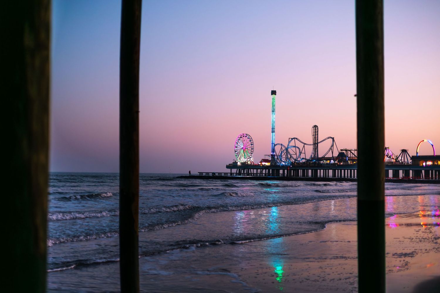 Colorful amusement park pier with Ferris wheel at sunset over calm ocean waves on a beach.
