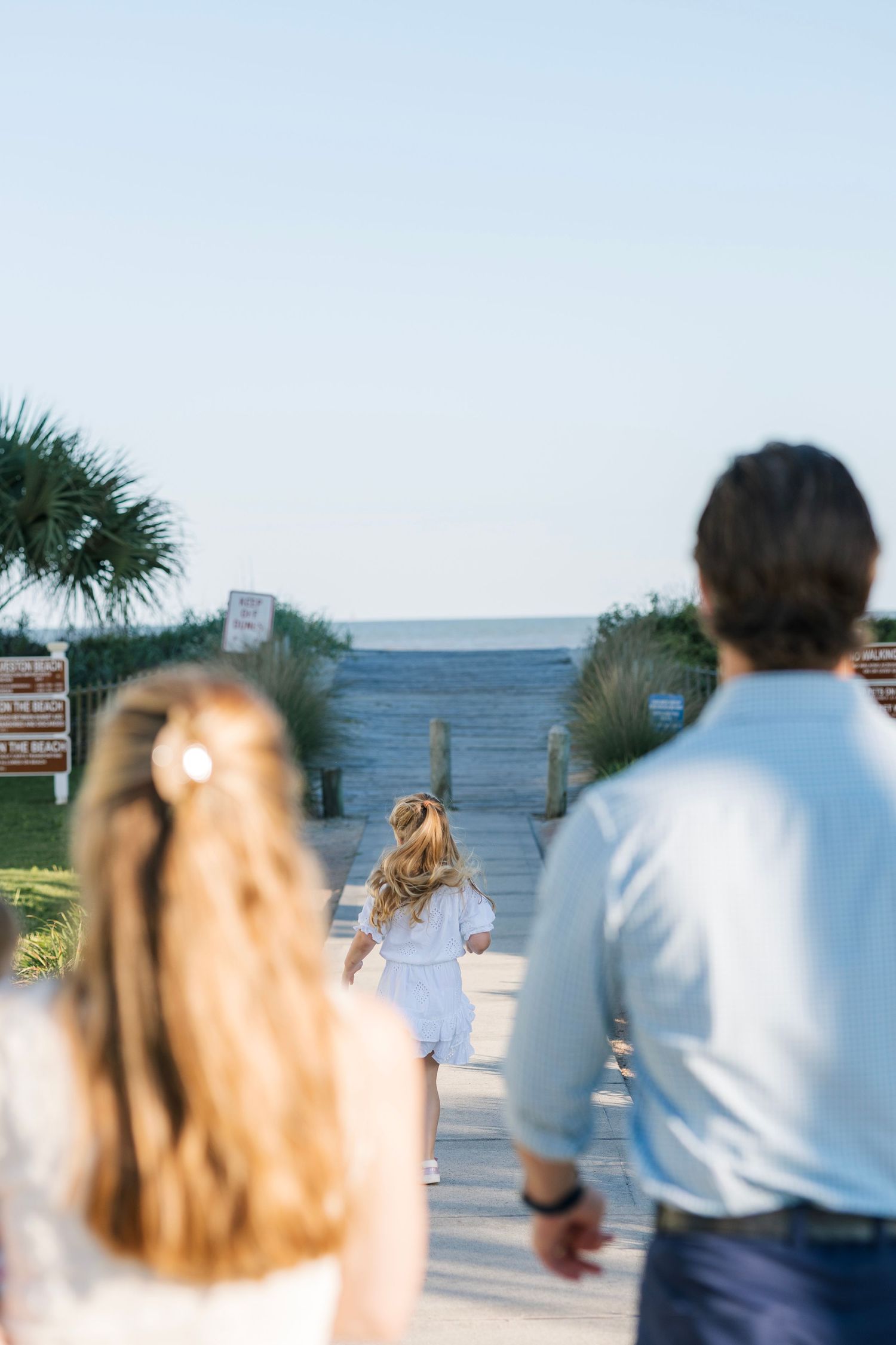 A family walks toward a beach boardwalk entrance surrounded by palm trees.