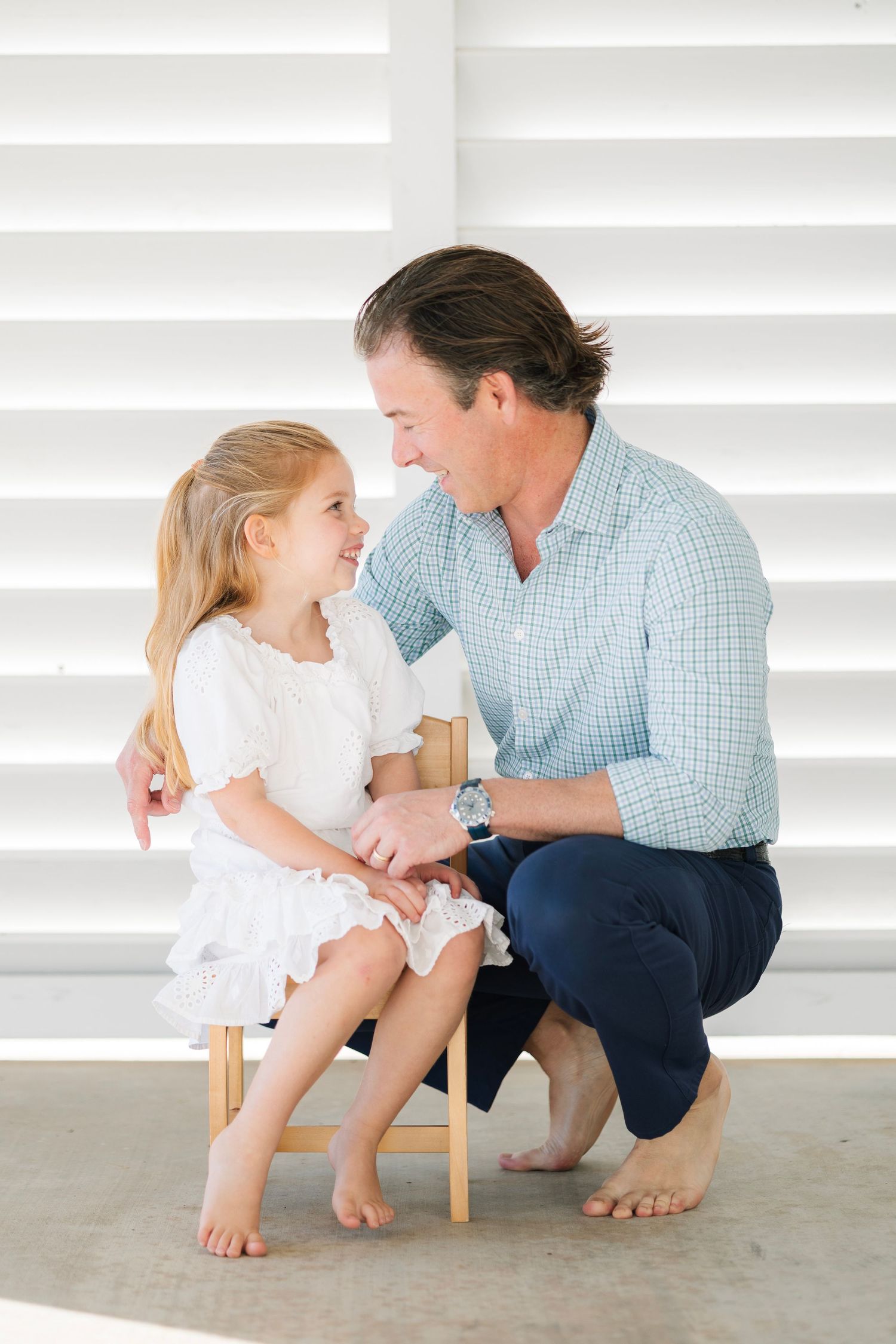A smiling man crouches beside a young girl in a white dress seated on a wooden chair against white shutters.