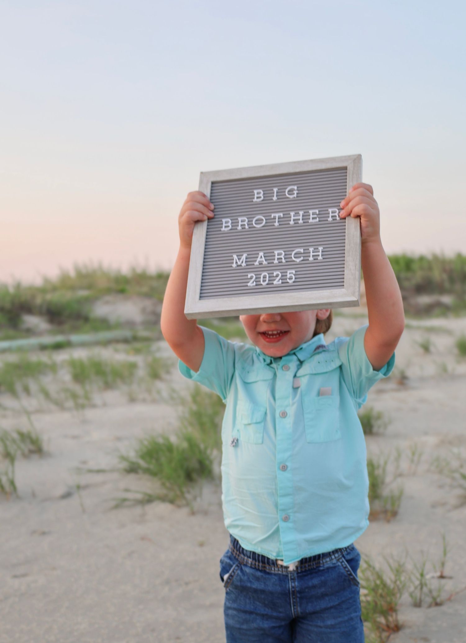 Child holding letterboard announcement on beach with big brother message.