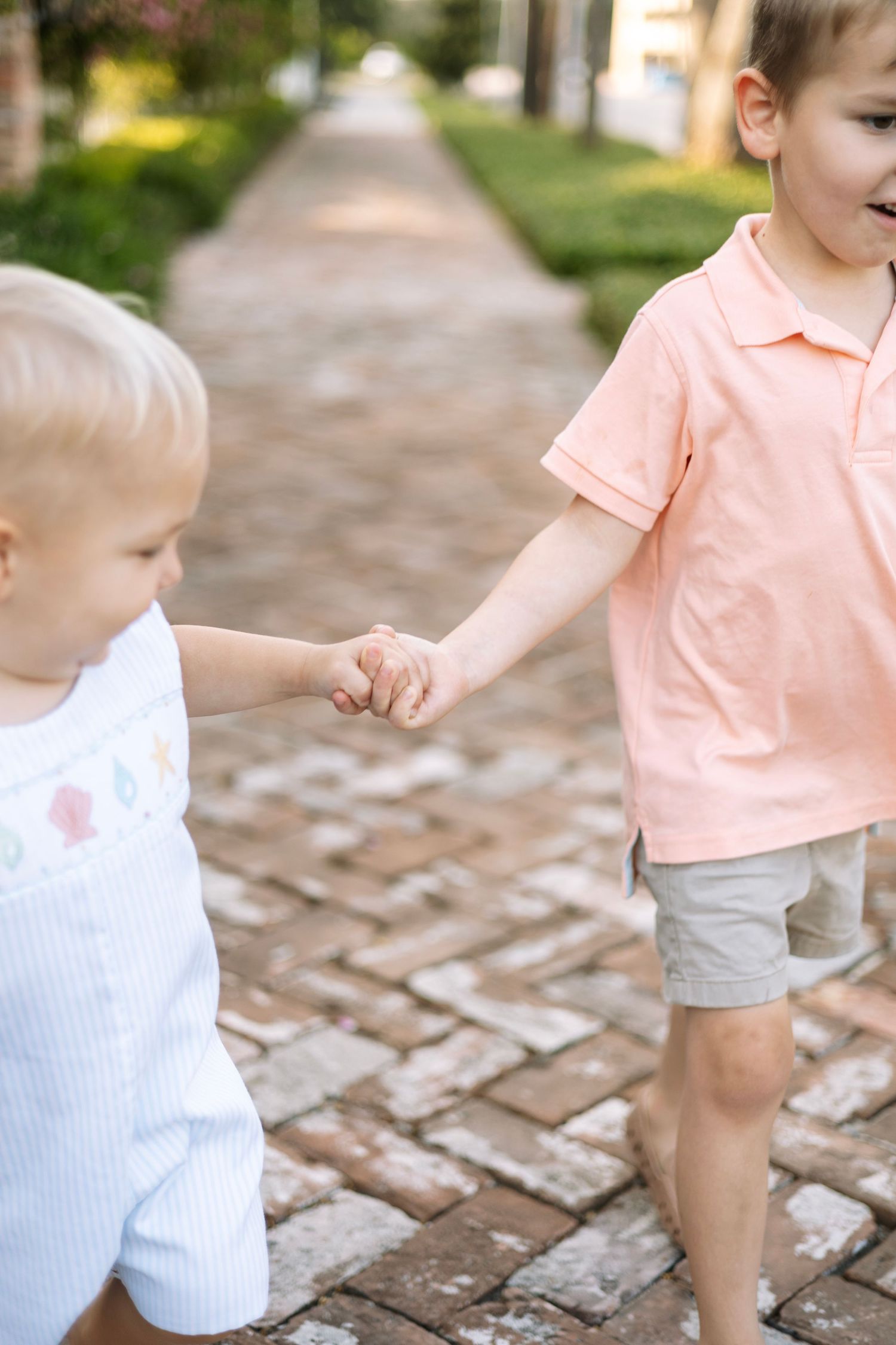 Two young children holding hands while walking on a brick pathway outdoors, wearing pastel summer outfits.