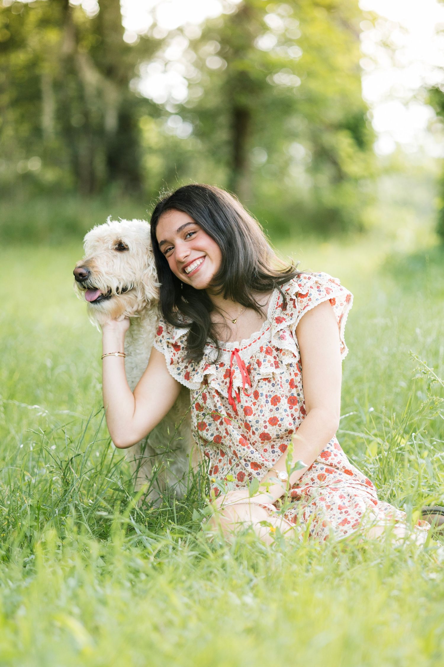 Smiling young woman in floral dress sitting in green grass outdoors, hugging a fluffy white dog on a sunny day.