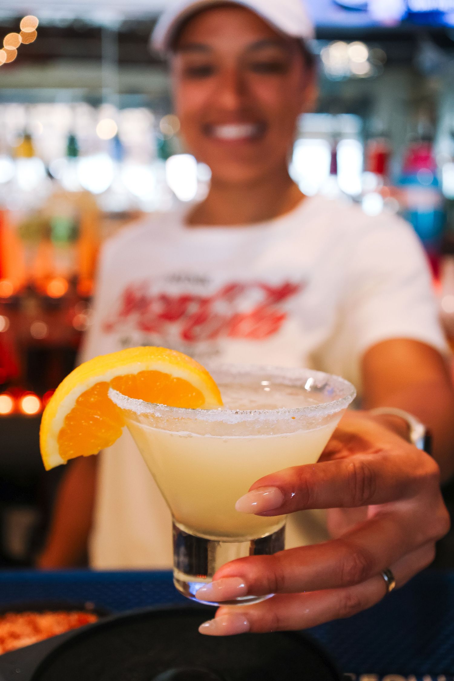 Smiling bartender holding a salted rim margarita garnished with an orange slice at a colorful bar.