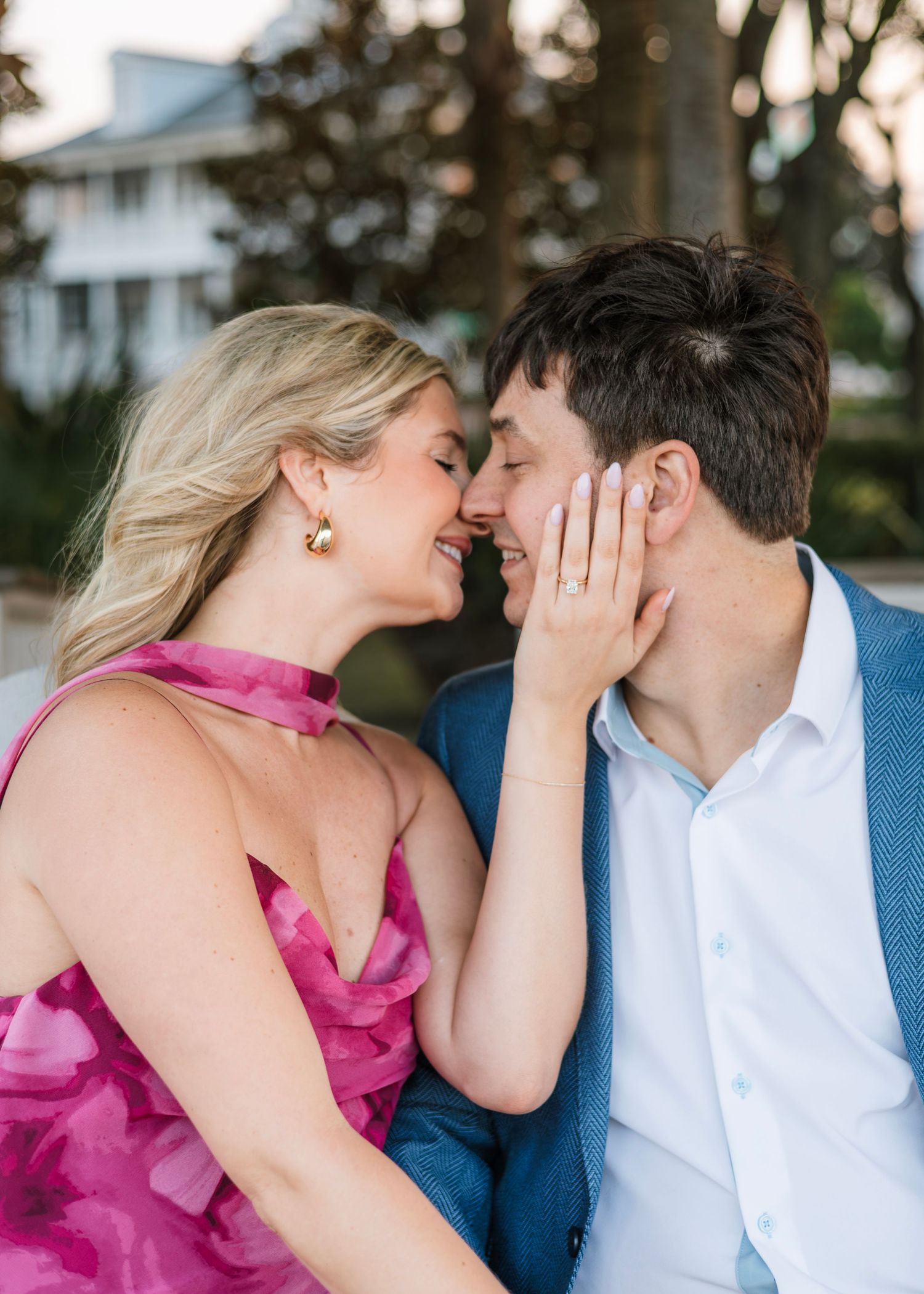 Couple in love almost kissing, woman in pink dress with engagement ring, man in blue suit, outdoor setting.
