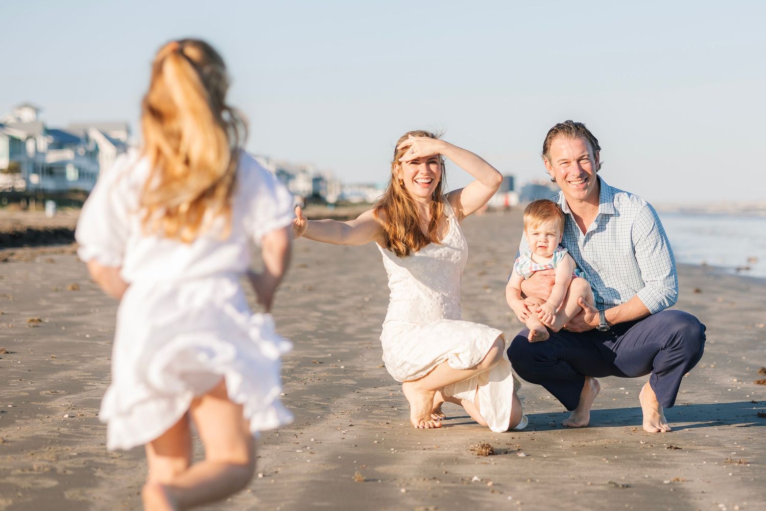 Family enjoying a sunny beach day, parents smiling as young child runs toward them on sandy shore.