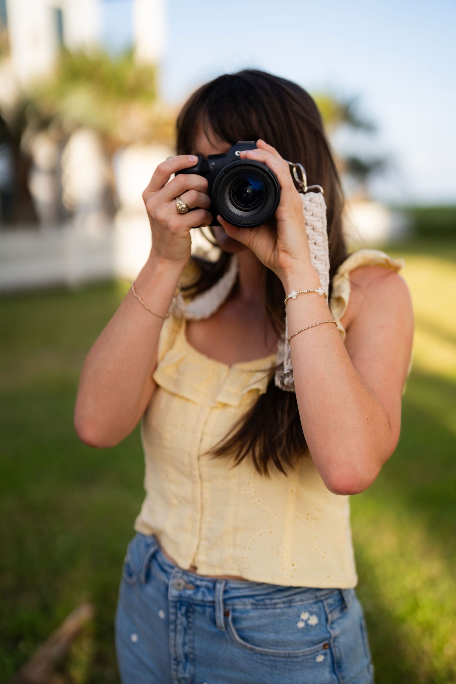 A photographer in a yellow top and jeans holds up a camera to take photos outdoors on a sunny day.
