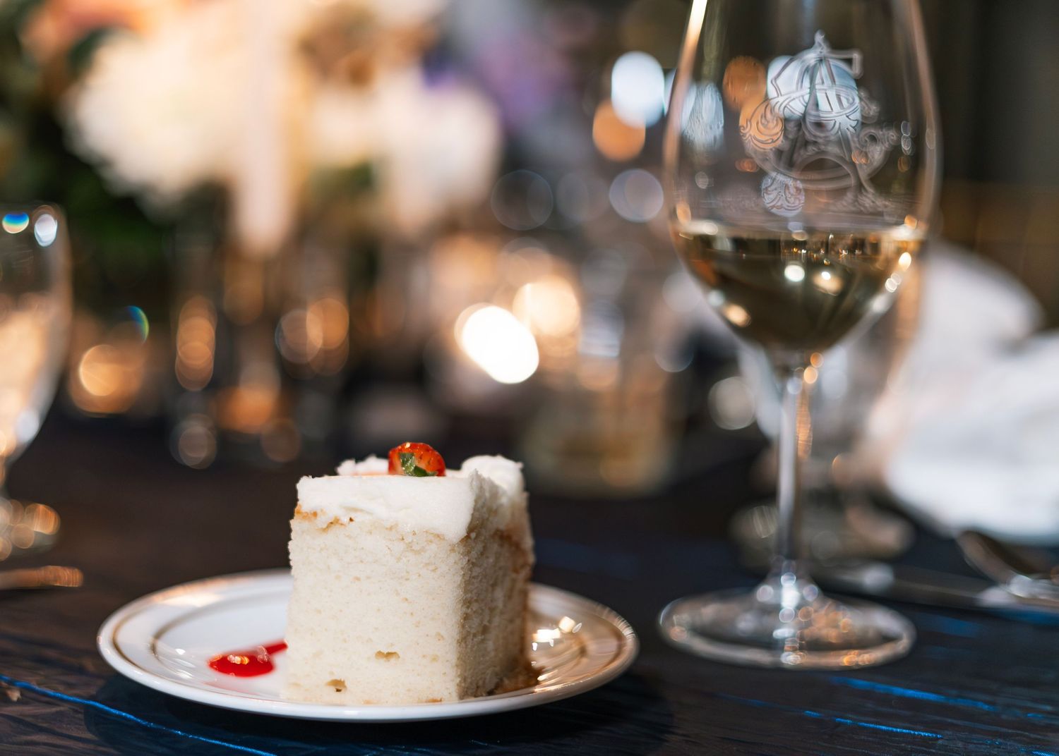 A slice of white cake with garnish served on a dark plate next to a glass of wine with bokeh lighting.