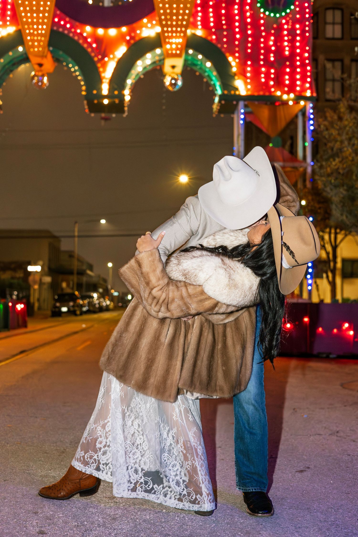 Couple in cowboy hats embracing on a festive night street with colorful lights overhead.