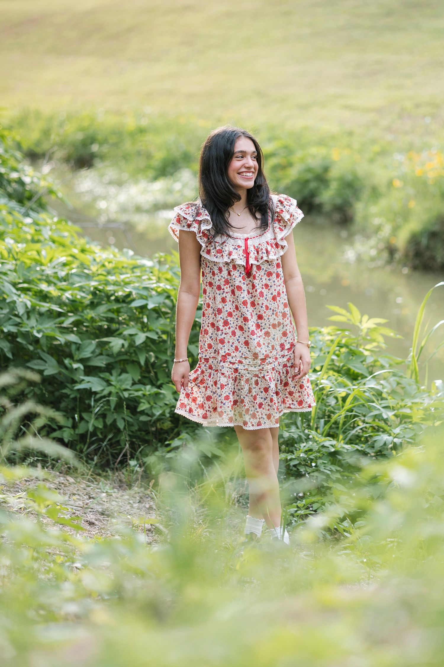 Young woman in floral dress smiling outdoors among lush green plants near a stream.