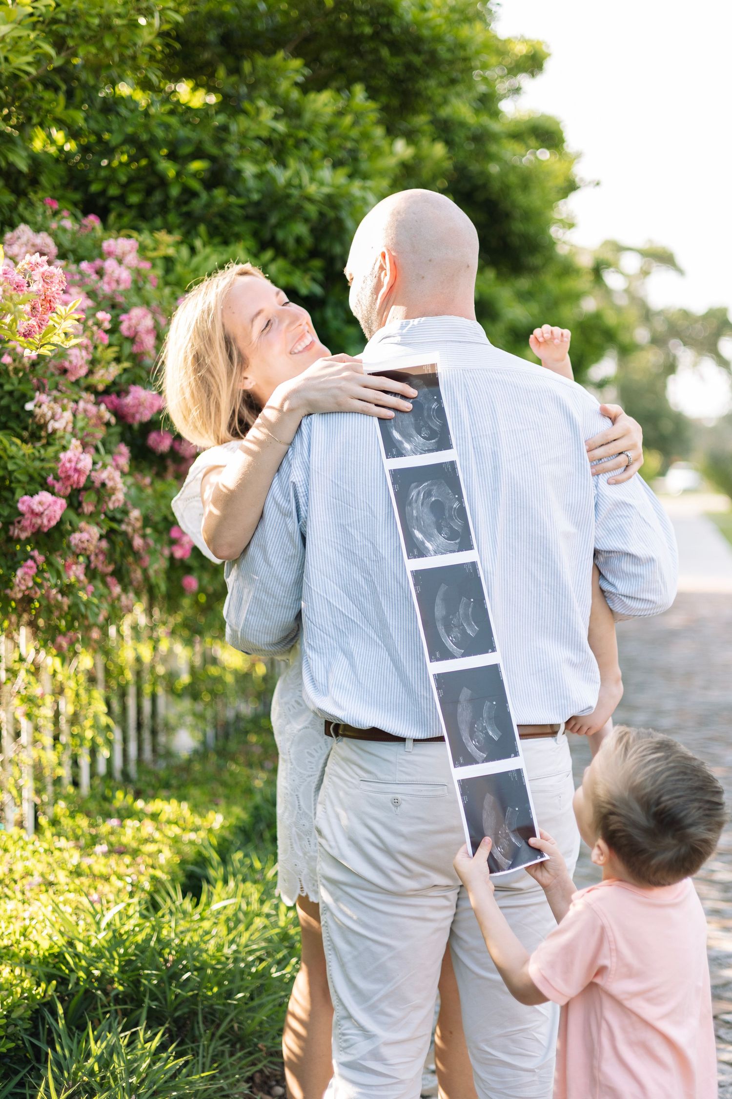 Happy couple embracing outdoors while child holds ultrasound photos, announcing pregnancy near blooming pink roses.