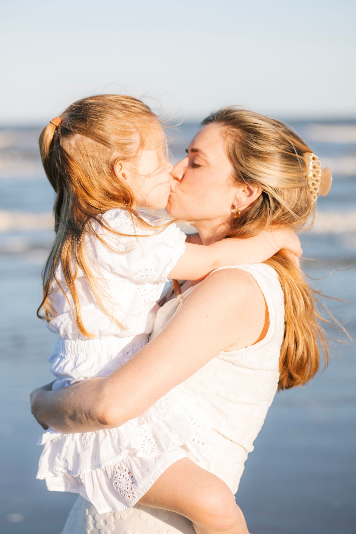 A mother and young daughter share a tender kiss on a sunny beach, both wearing white dresses.