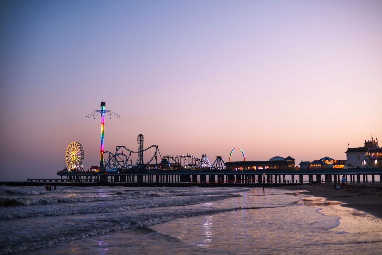 Colorful amusement park rides and Ferris wheel light up a beachside pier at dusk with pastel sunset skies.