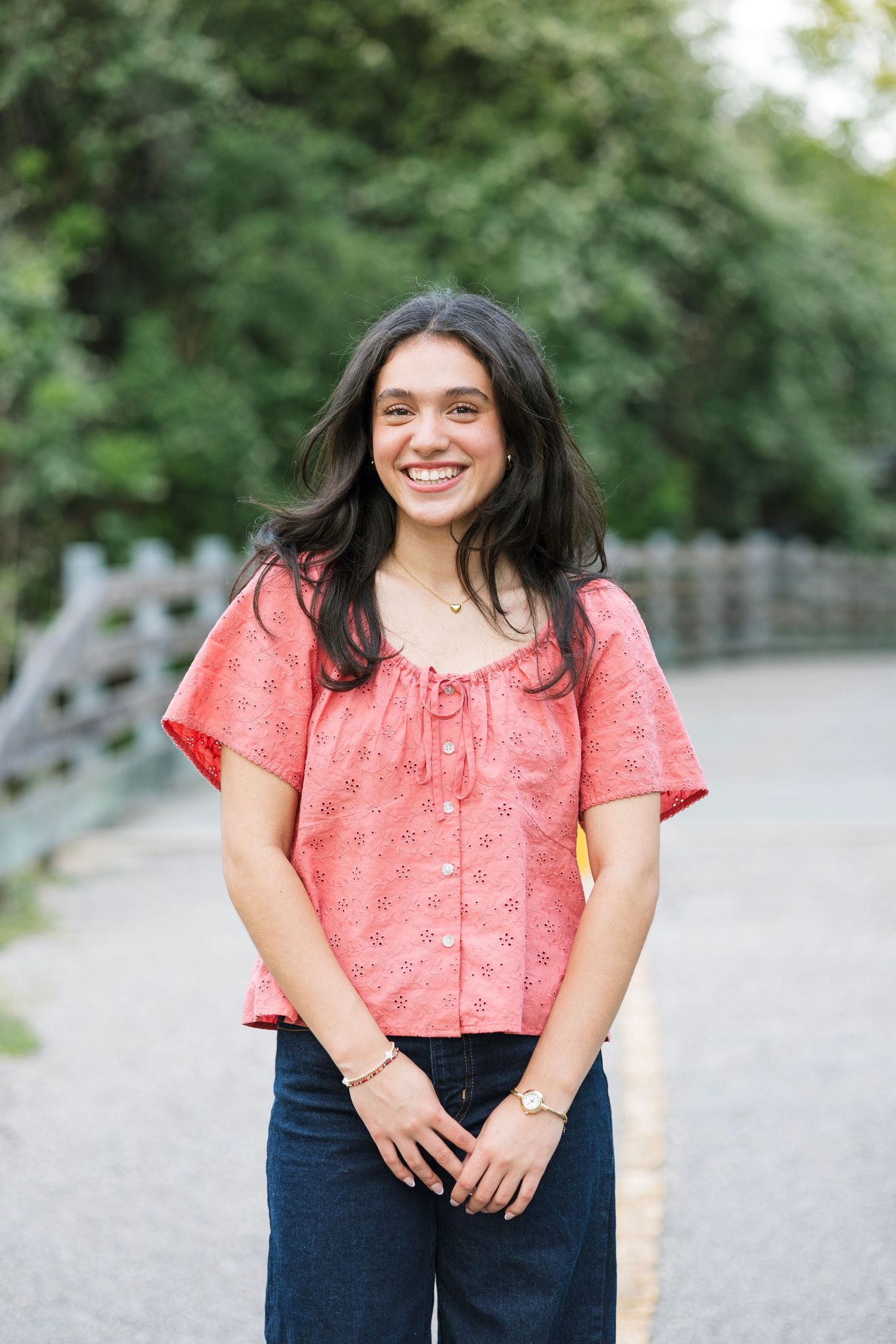 Smiling young woman in pink eyelet top and jeans standing on outdoor path surrounded by trees.