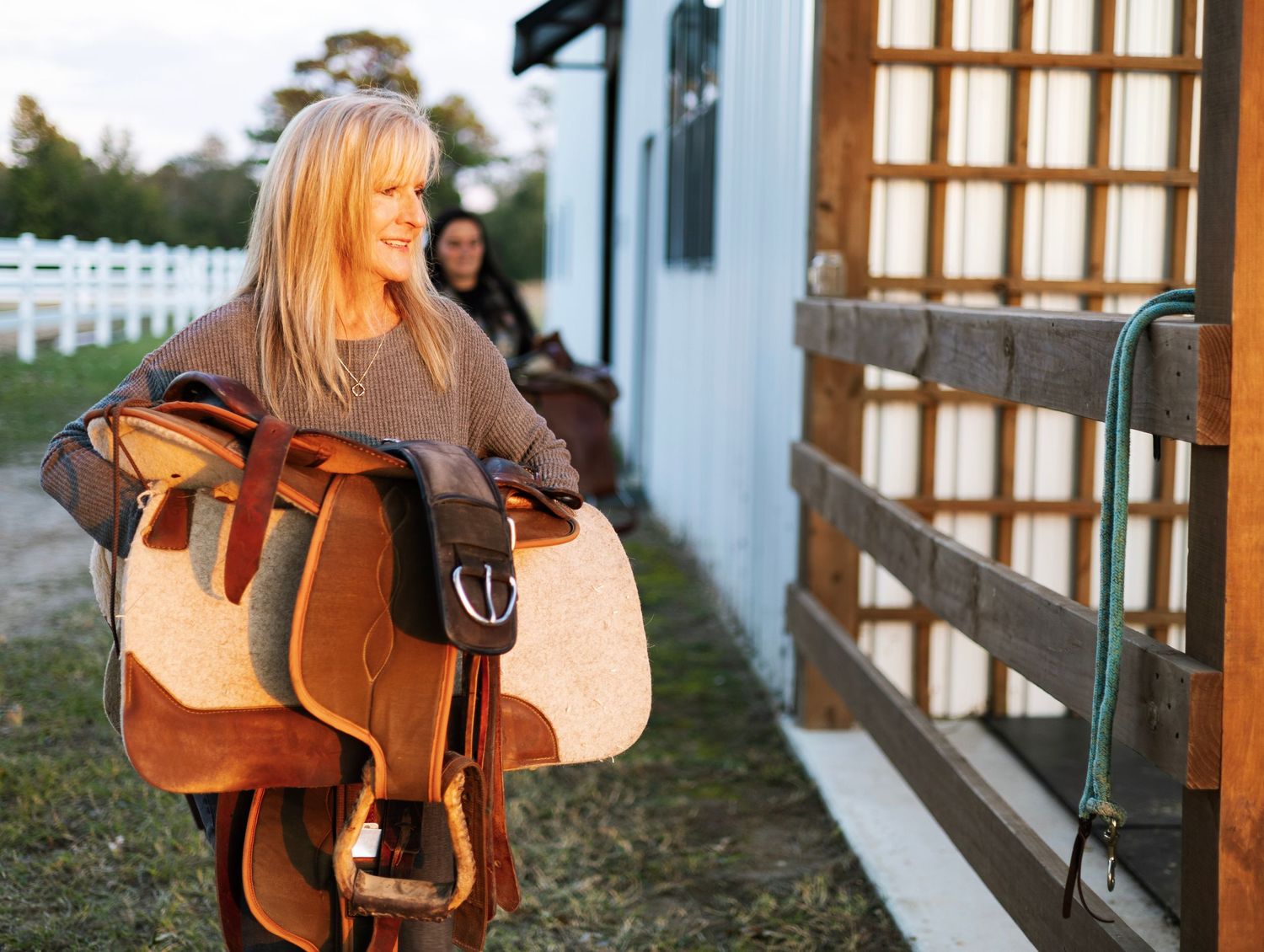 Blonde woman carrying a Western saddle outside a white stable building at a horse ranch.