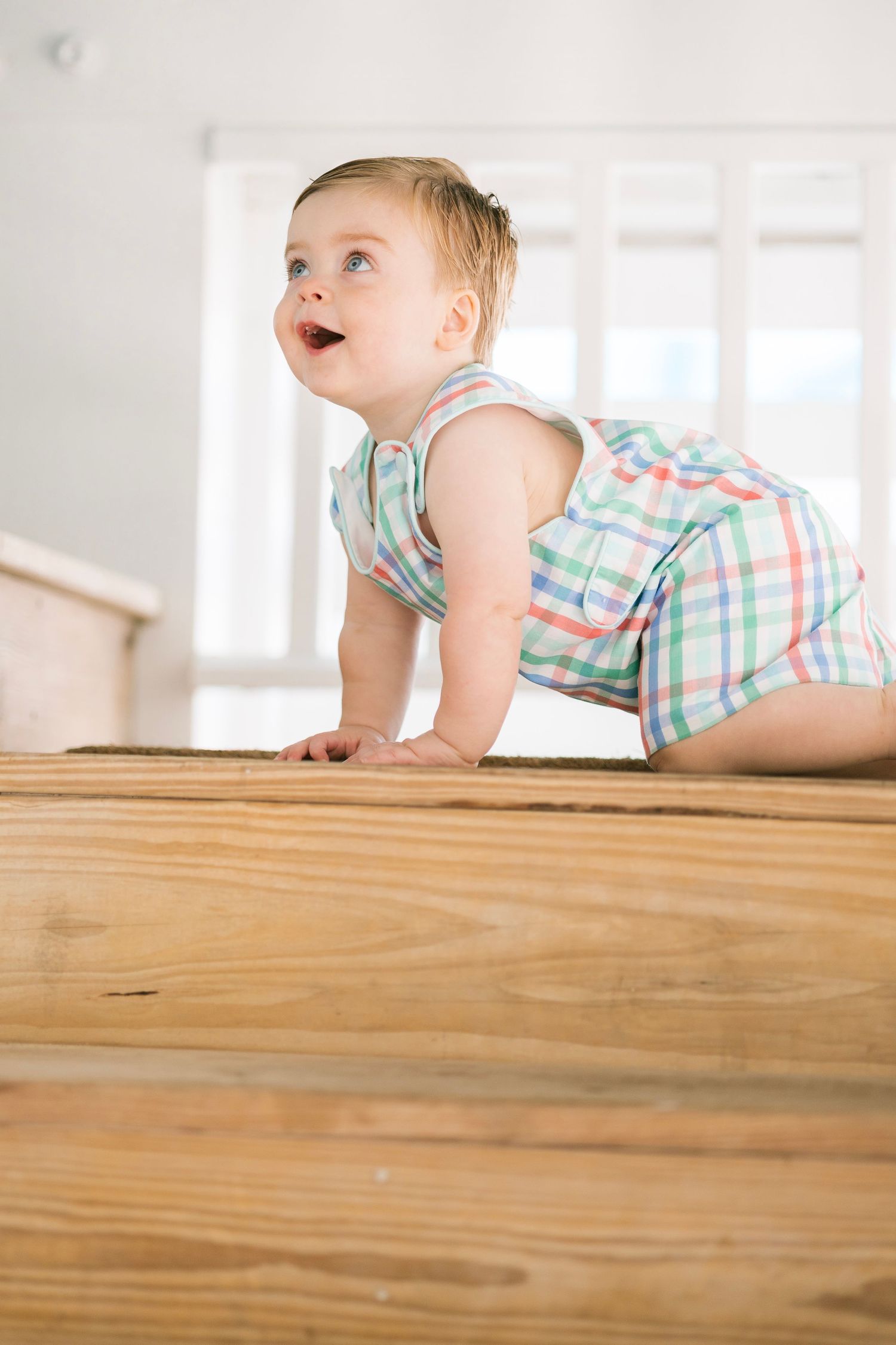 Curious baby in plaid romper crawling on hardwood floor, looking up with a happy expression.