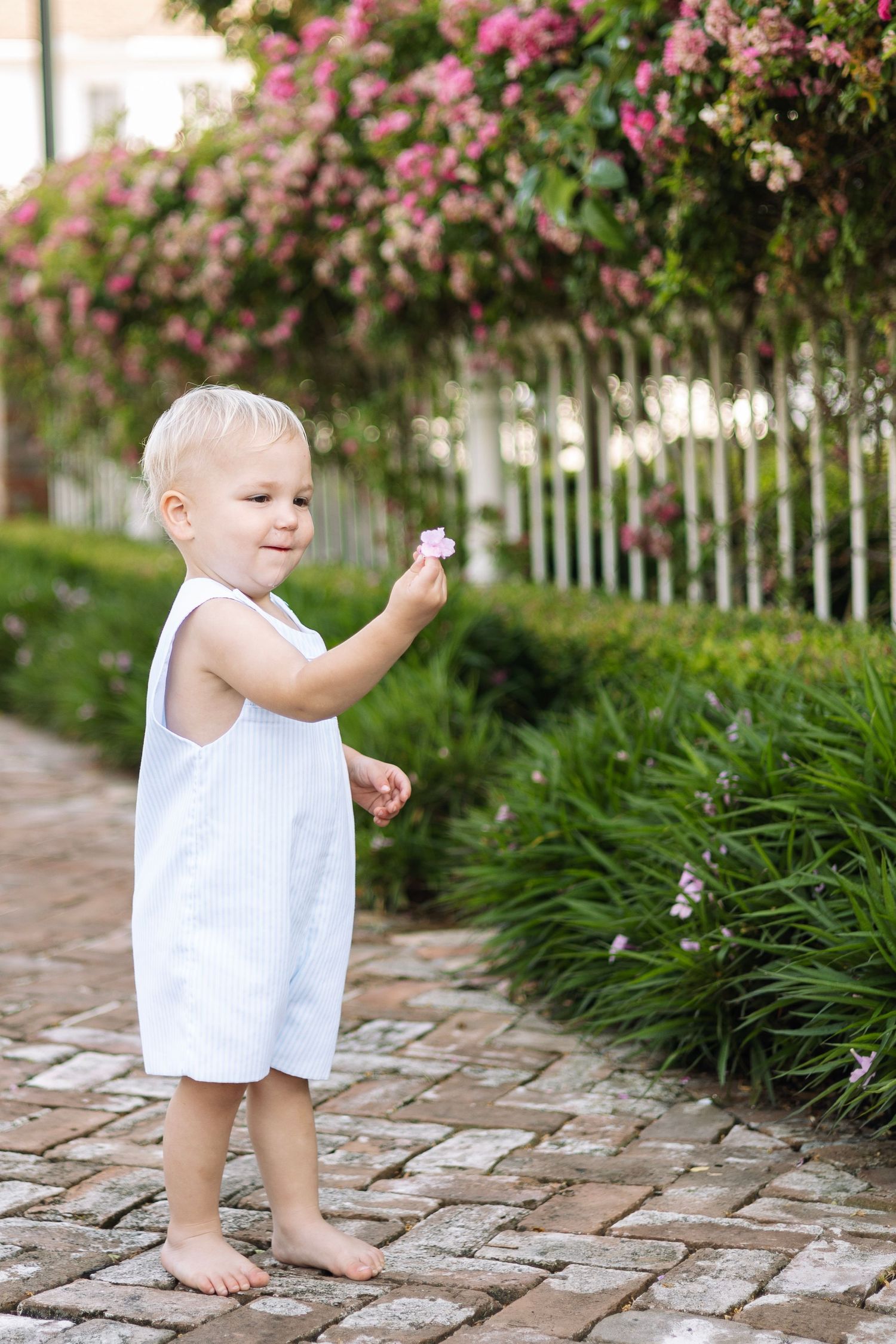 Toddler in white dress holding a pink flower, standing barefoot on brick path with blooming garden and white fence.