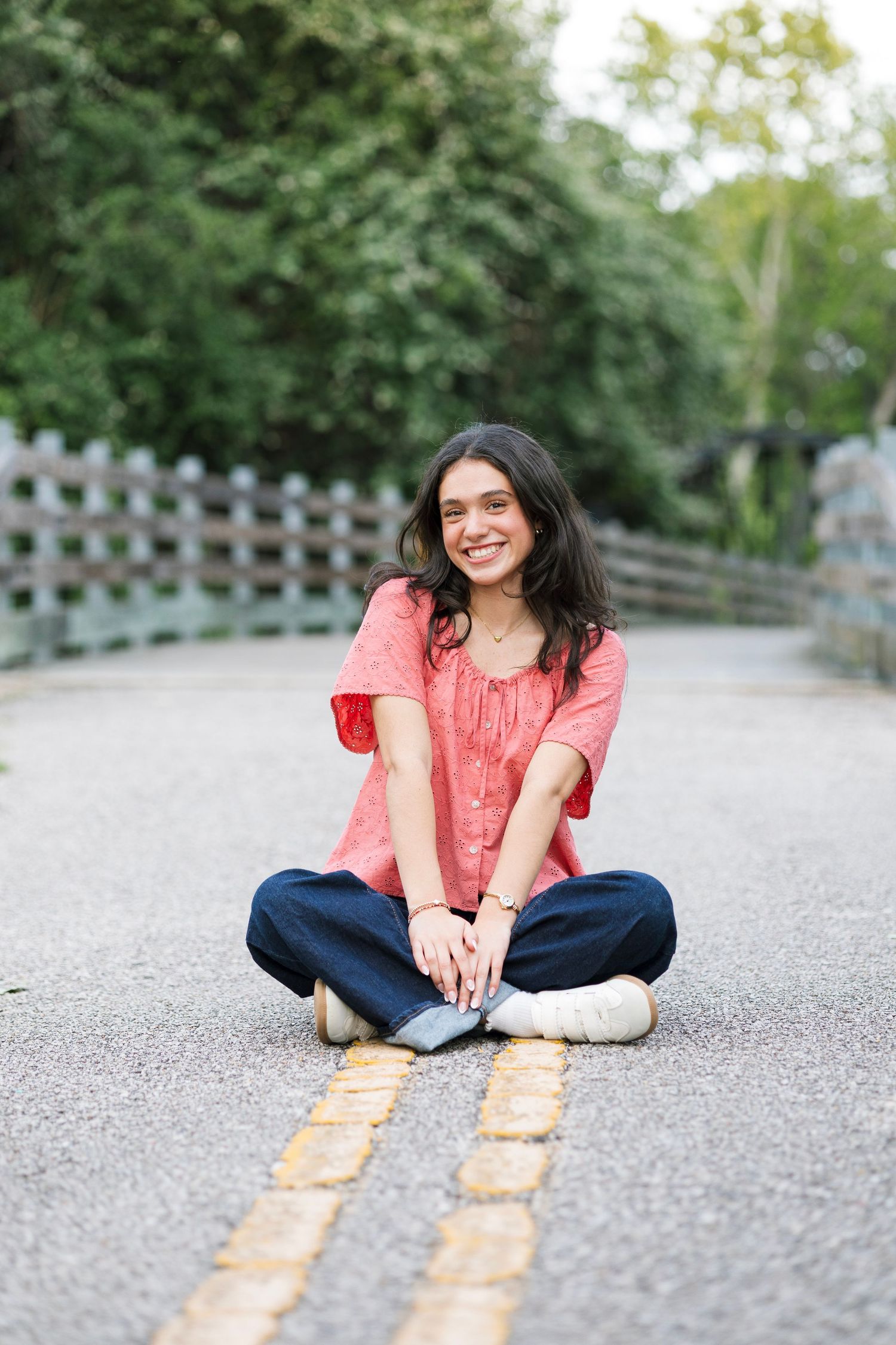 Smiling young woman in red top and jeans sitting cross-legged on a road in a park setting.