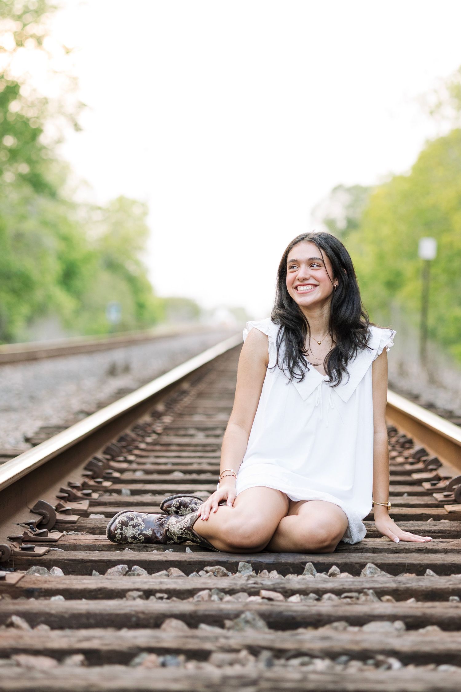 Young woman in white dress sitting cross-legged on railroad tracks, smiling joyfully, surrounded by lush green trees.