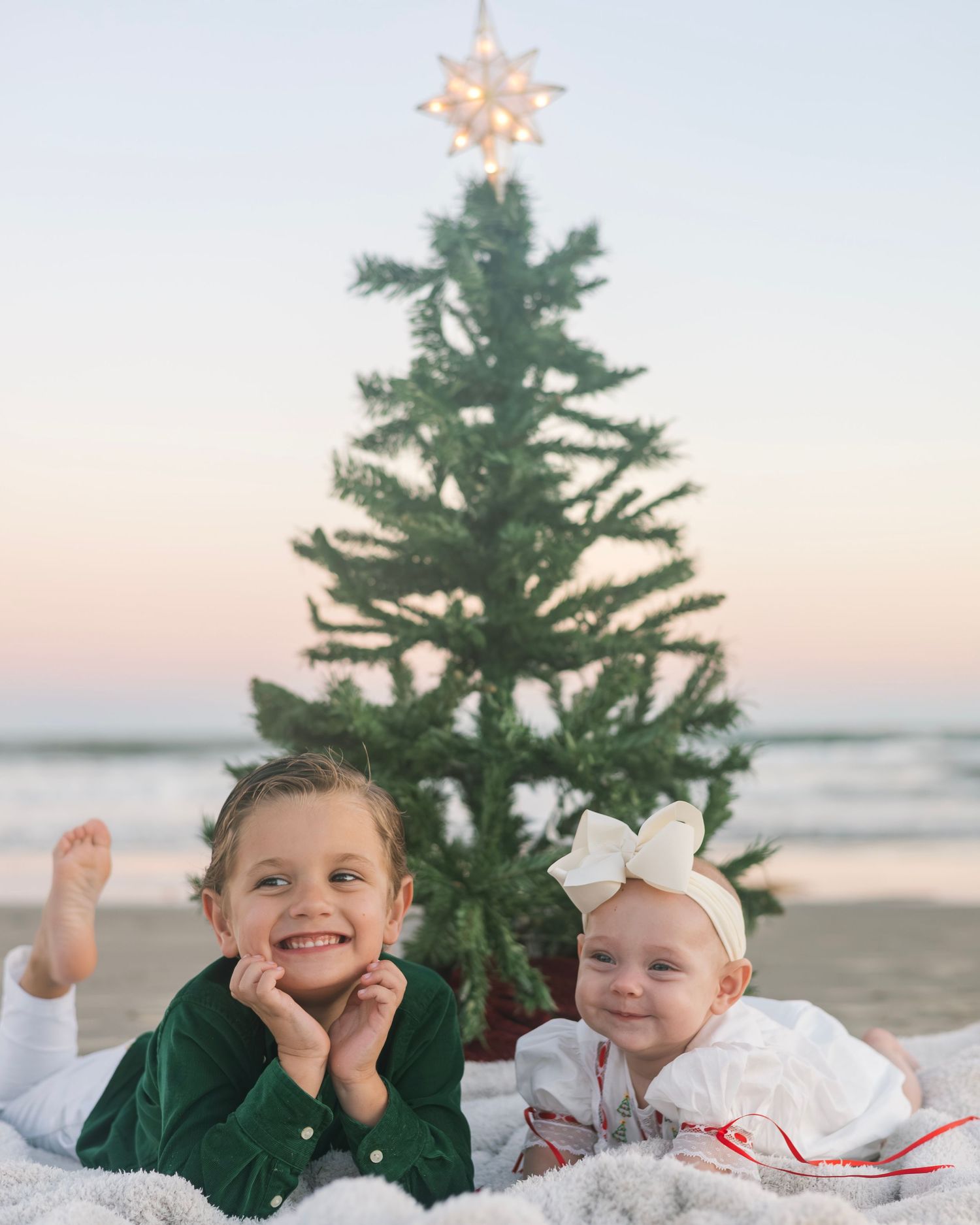 Children posing with a small Christmas tree on a sandy beach during golden hour.