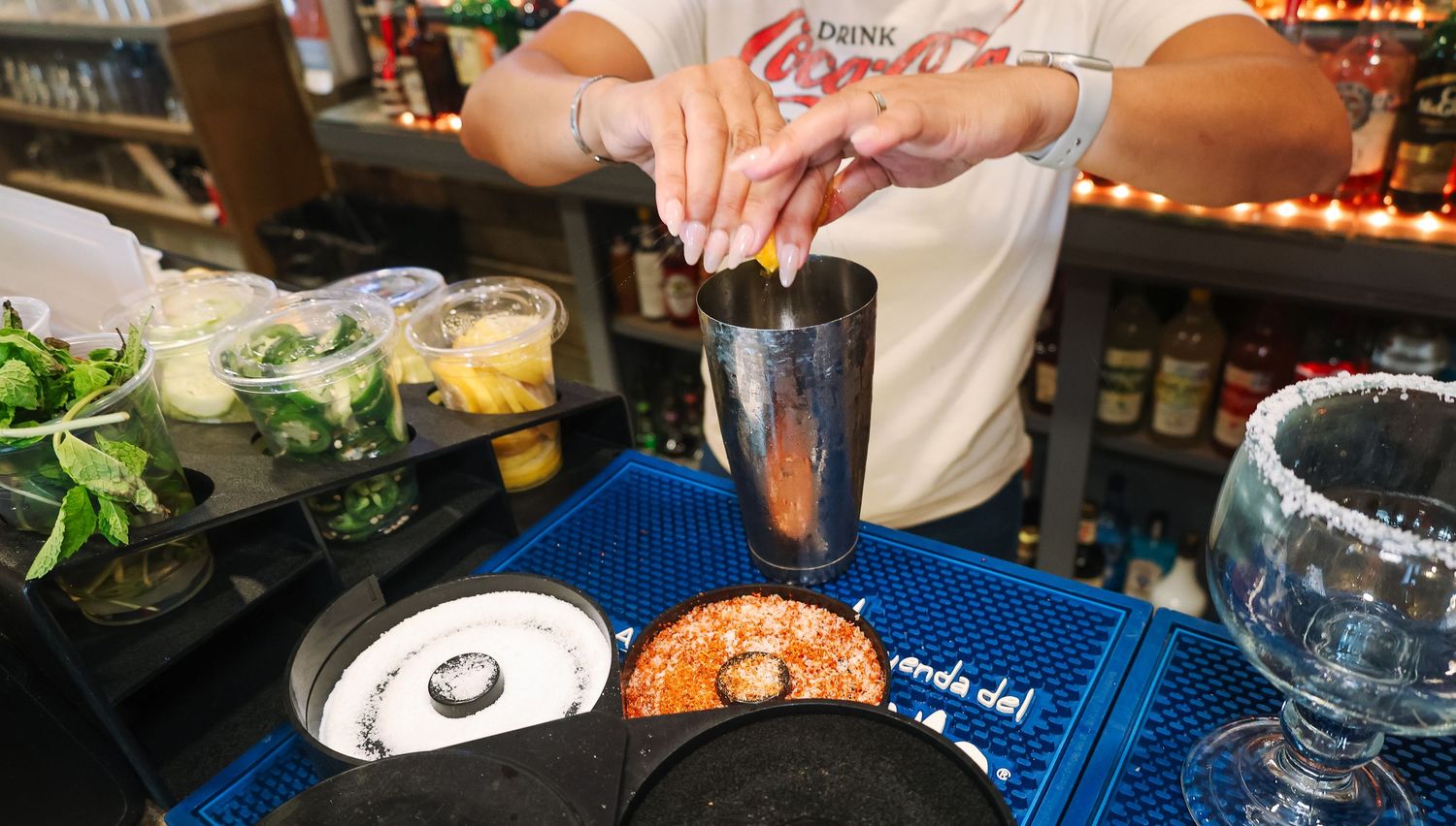 Bartender shaking cocktail in metal shaker at a bar with fresh ingredients and bottles in background.