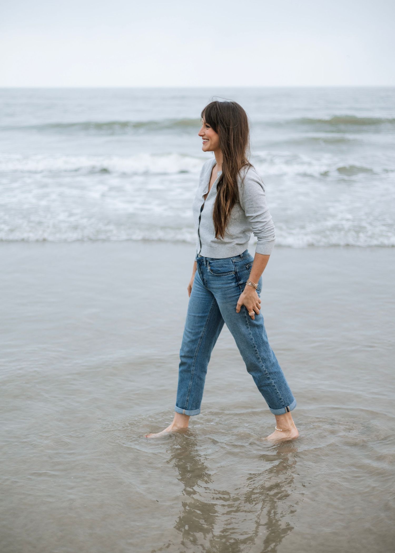 Woman walking barefoot on a beach in blue jeans and gray cardigan, smiling with ocean waves behind her.