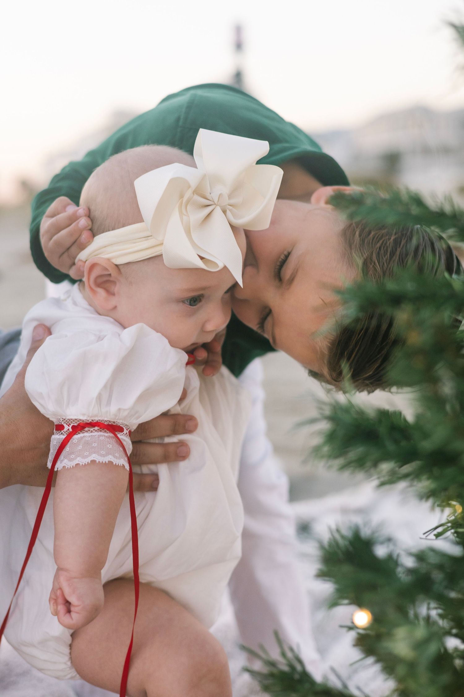 A series of photos showing a baby in a white dress with red accents near evergreen branches.