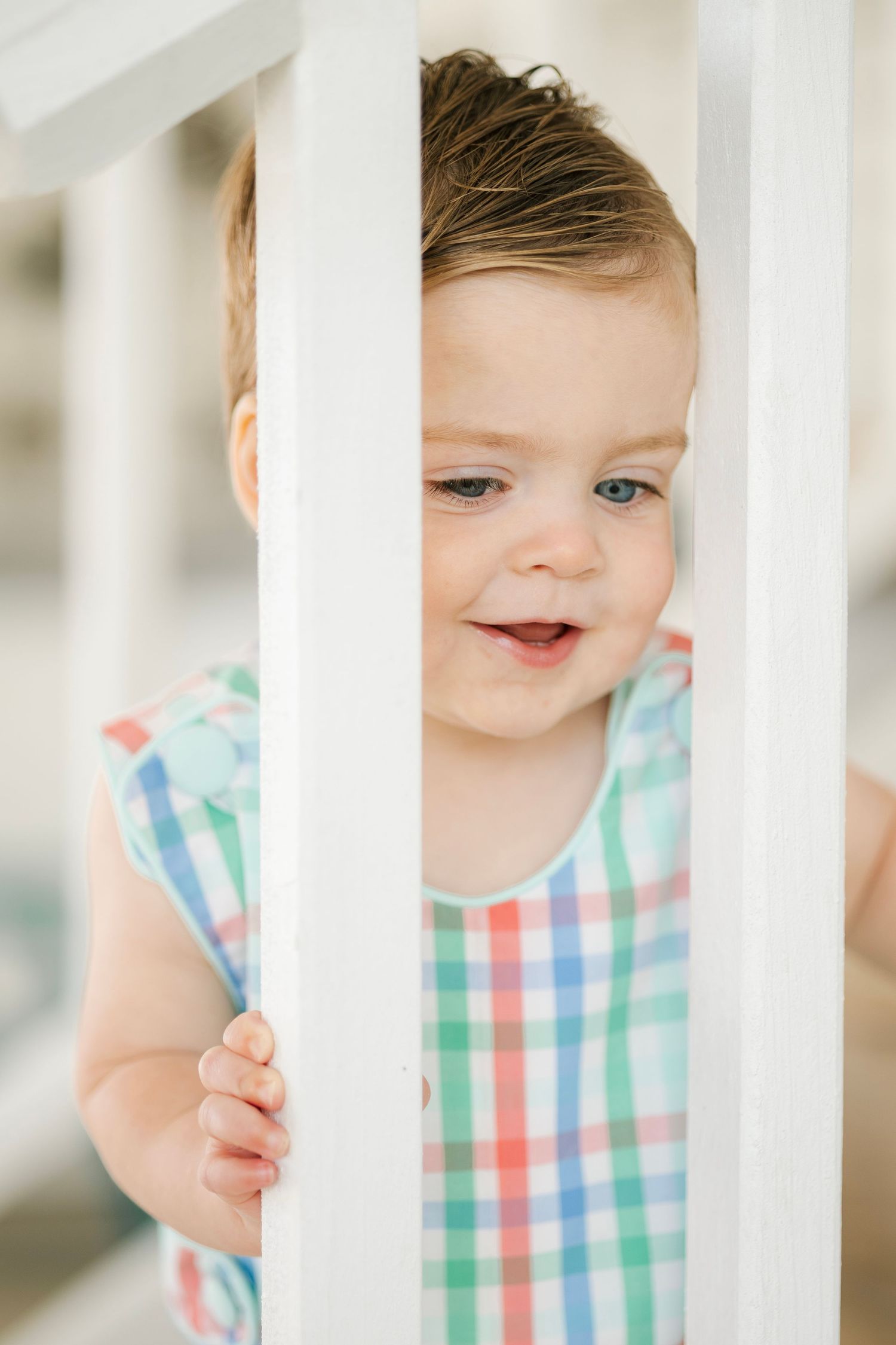 Smiling baby with blue eyes peering through white crib rails wearing colorful plaid romper.