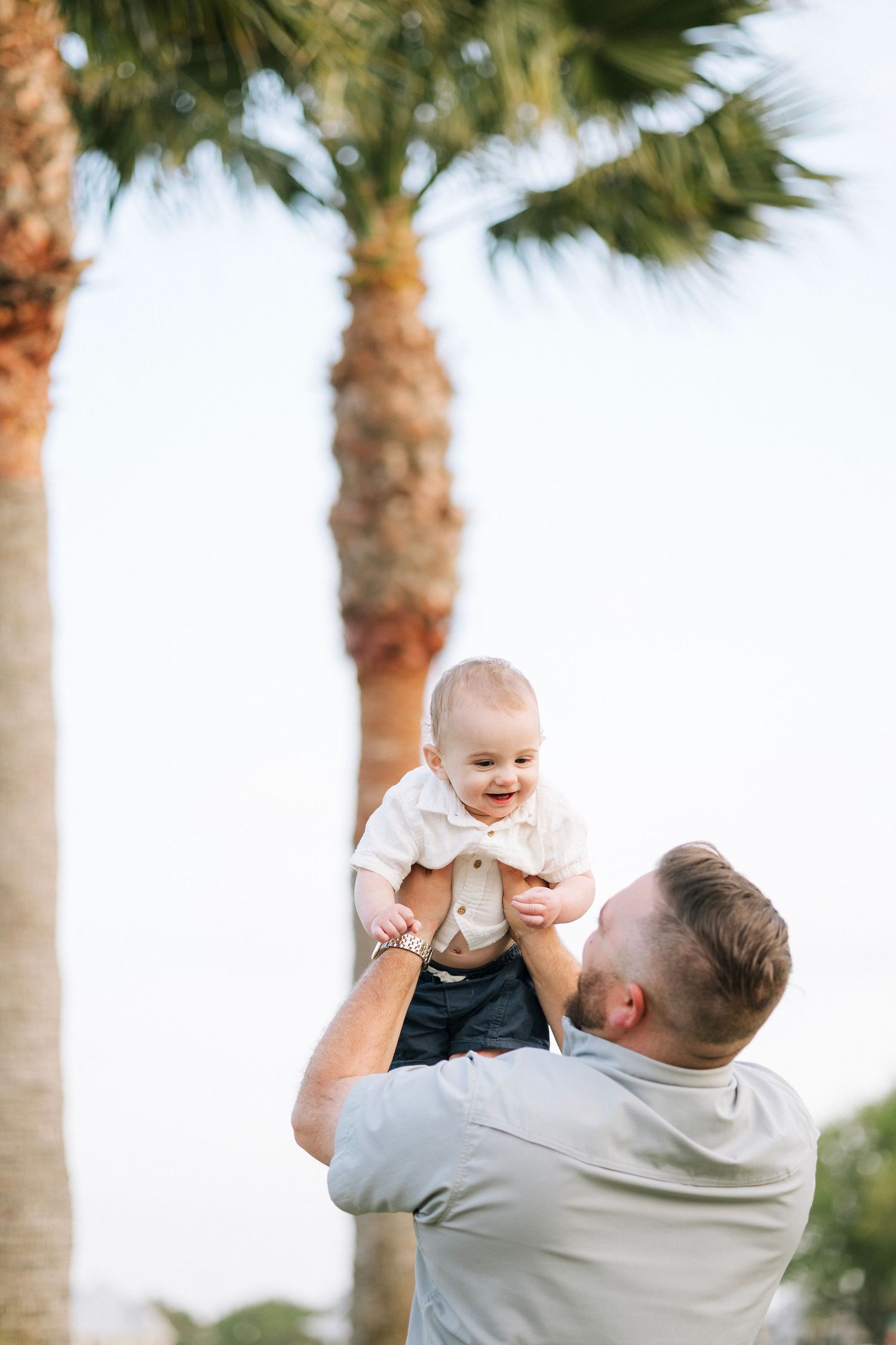 Father joyfully lifts smiling baby in the air outdoors near tropical palm trees on a bright sunny day.