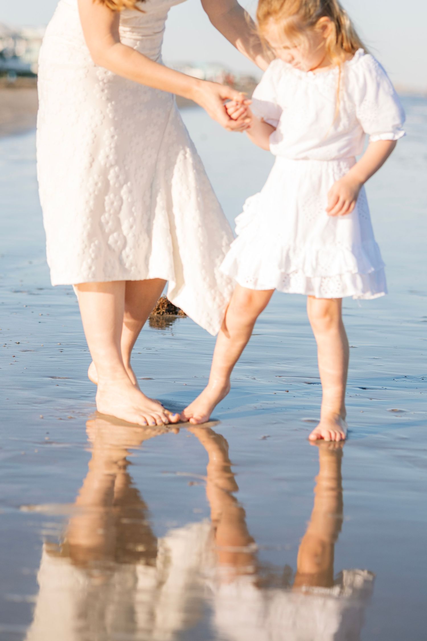 Mother and daughter in white dresses dancing barefoot on a reflective wet beach shore.