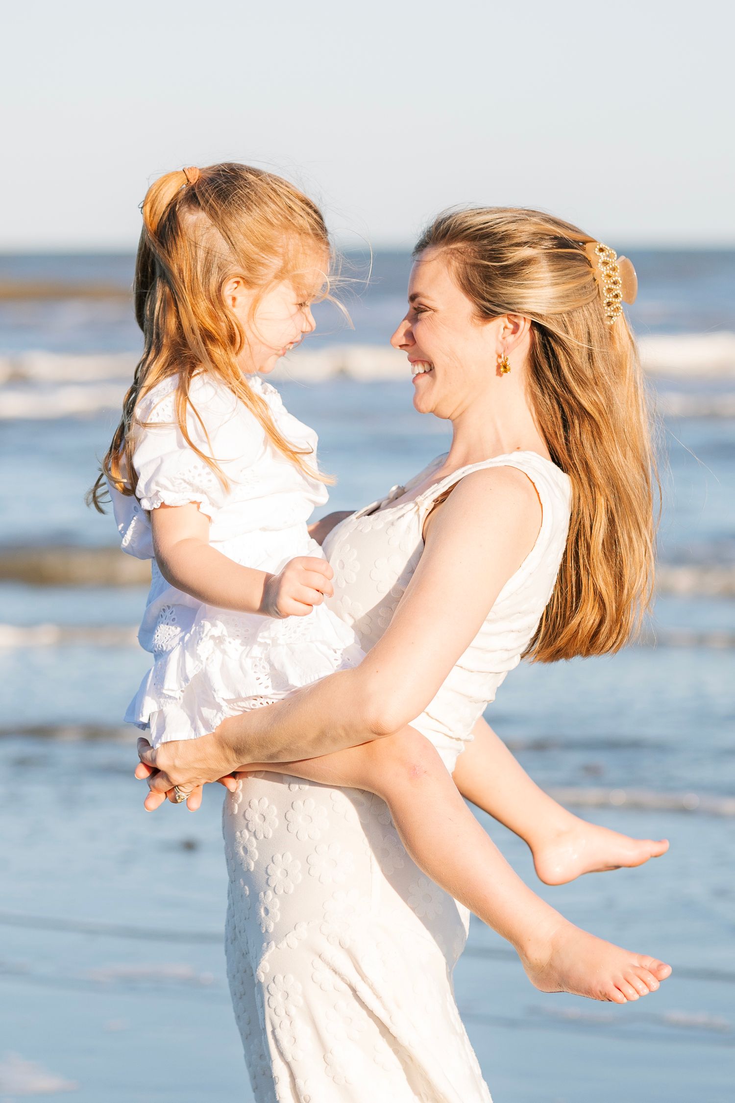 A joyful mother in a white dress holds her laughing young daughter on a sunny beach with ocean waves behind them.