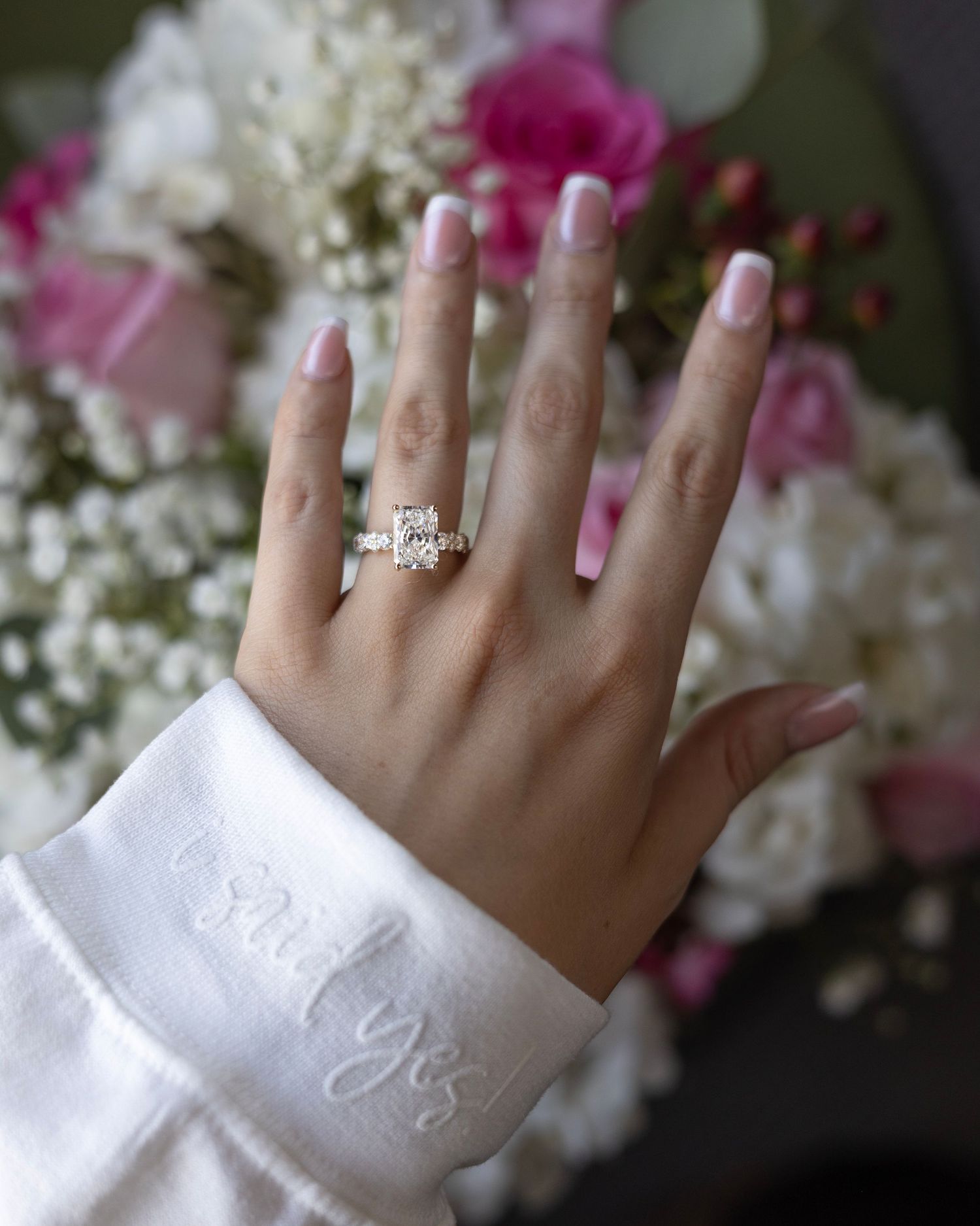 Close up of a hand wearing an engagement ring with white flowers in the background.
