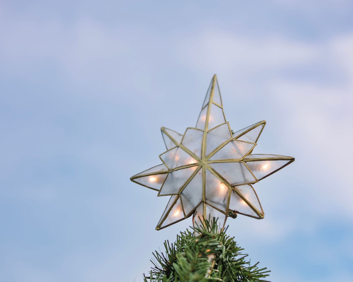 A silver star-shaped Christmas tree topper is photographed against a bright blue sky with wispy clouds.