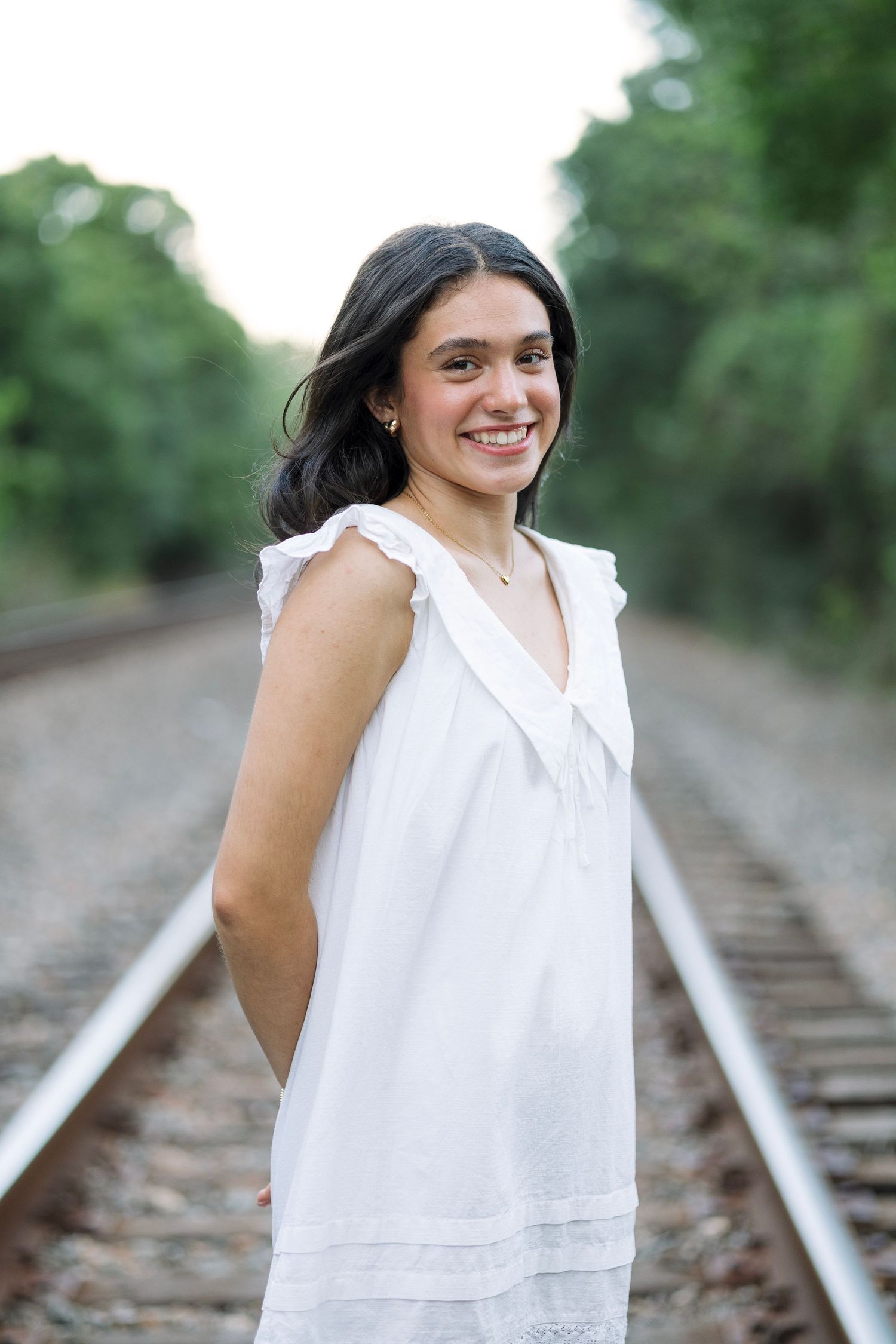 Smiling young woman in white dress standing on railroad tracks surrounded by lush green trees.