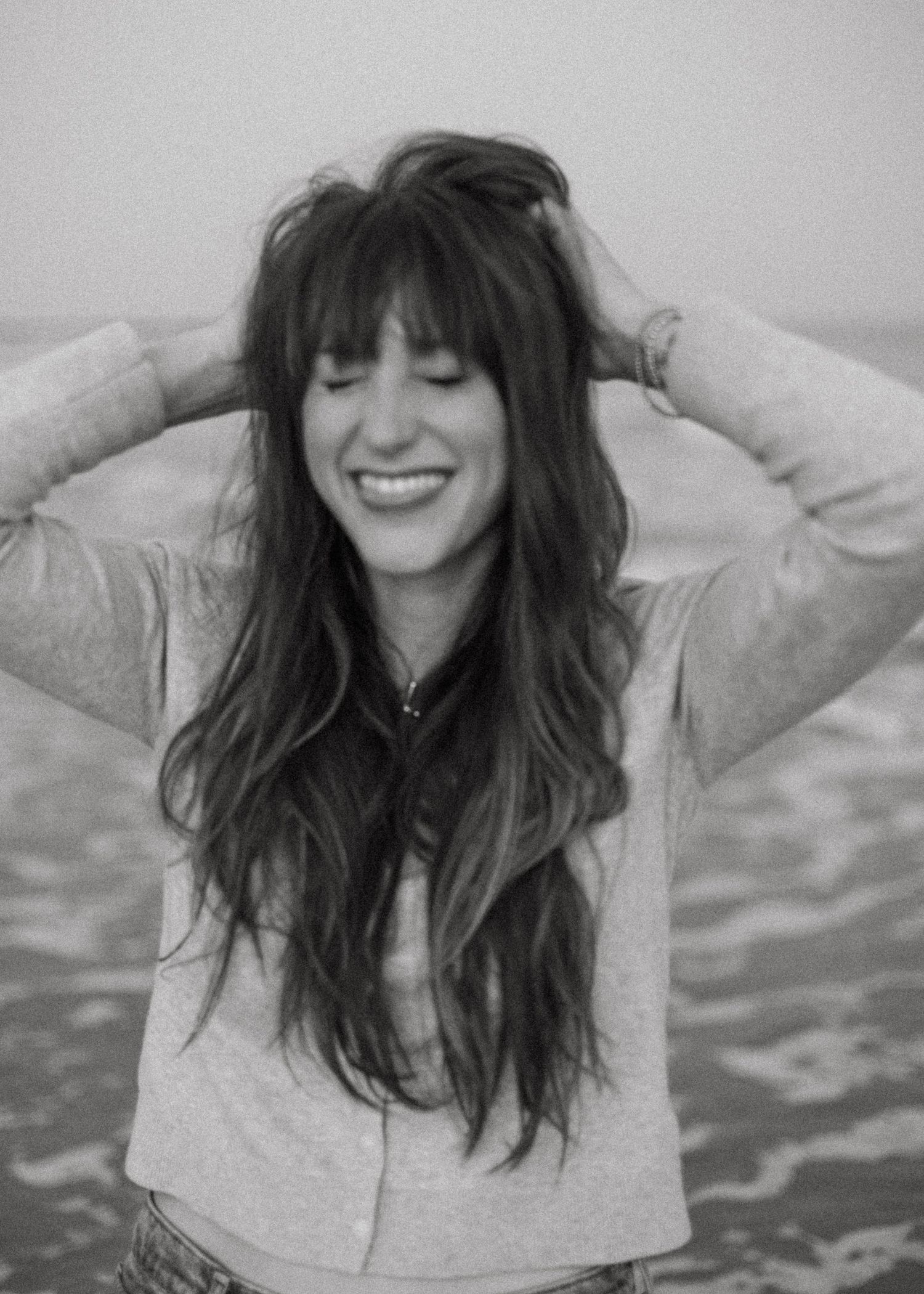 Black and white photo of a smiling woman with long wavy hair and bangs, standing near the ocean.