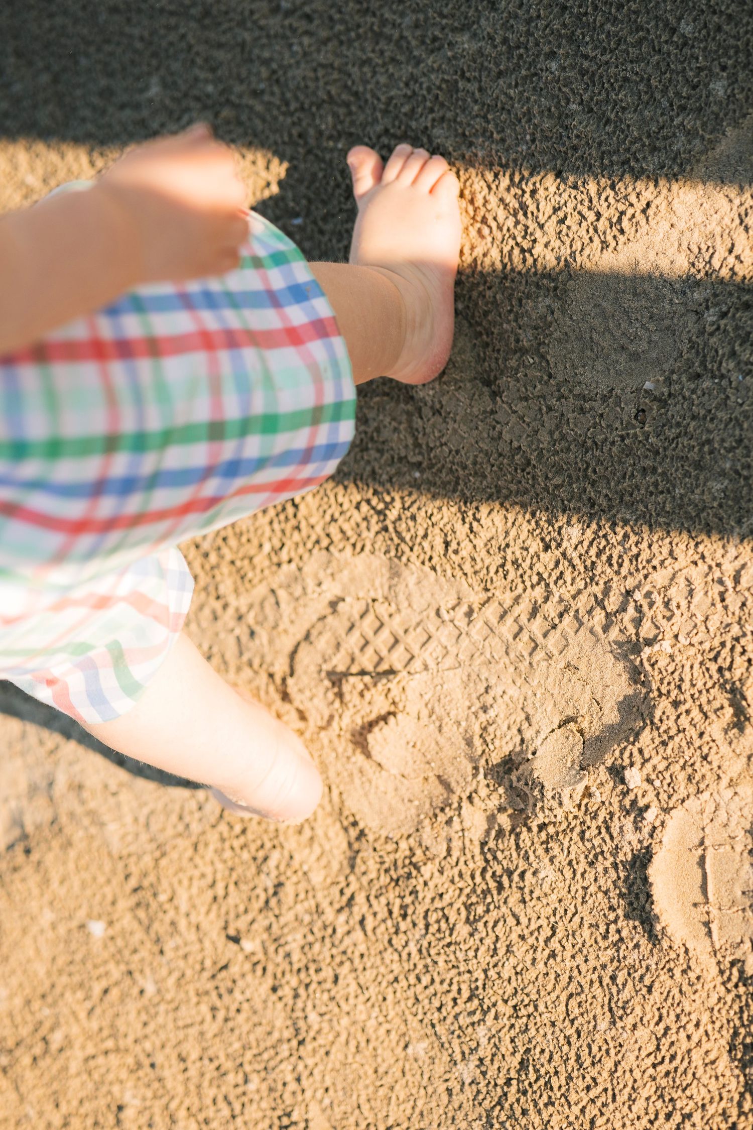 Child's bare feet standing in sandy beach with adult feet nearby, wearing colorful plaid shorts on sunny day.