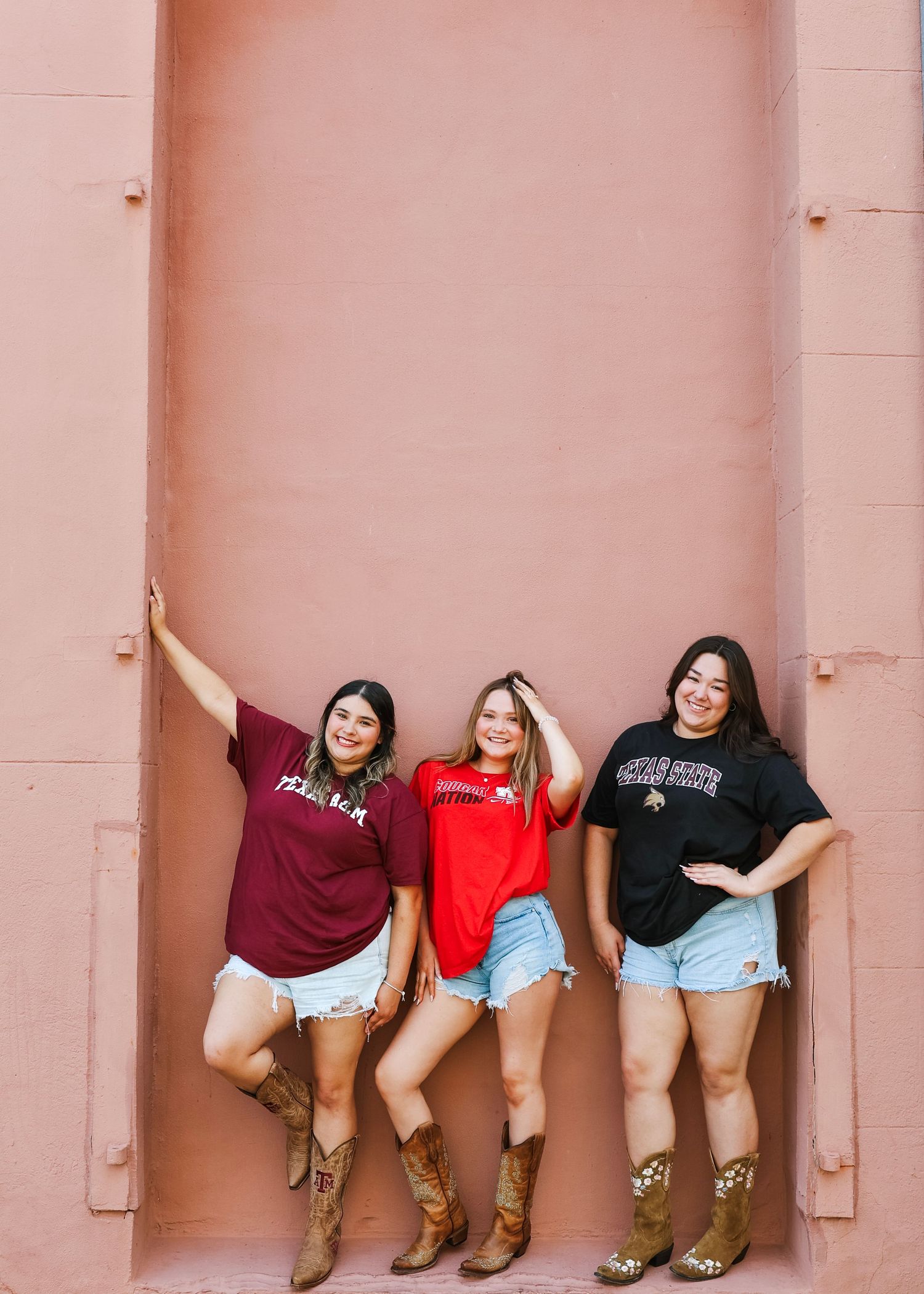 Friends posing playfully against a pink wall wearing casual t-shirts and denim shorts.