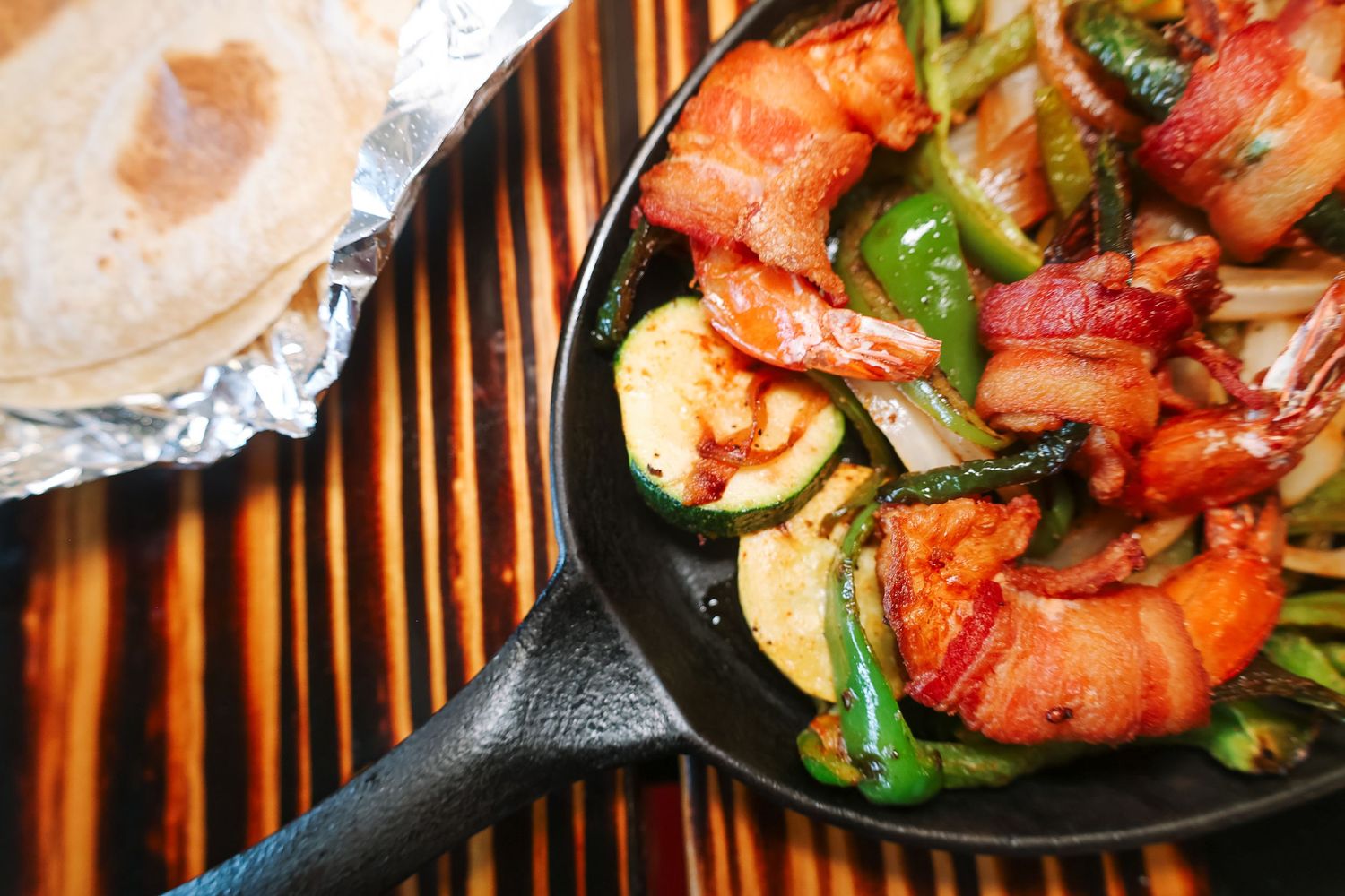 Cast iron skillet with bacon-wrapped shrimp, zucchini, and green peppers, served with warm tortillas in foil.