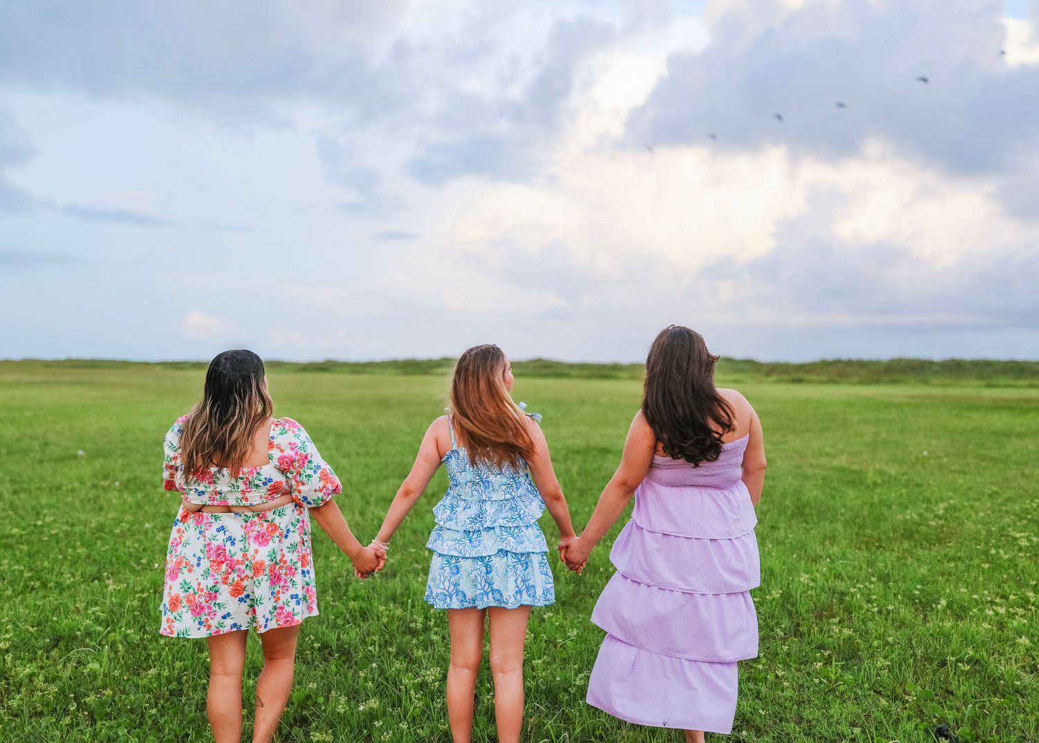 Three friends in summer dresses hold hands while walking through green field.
