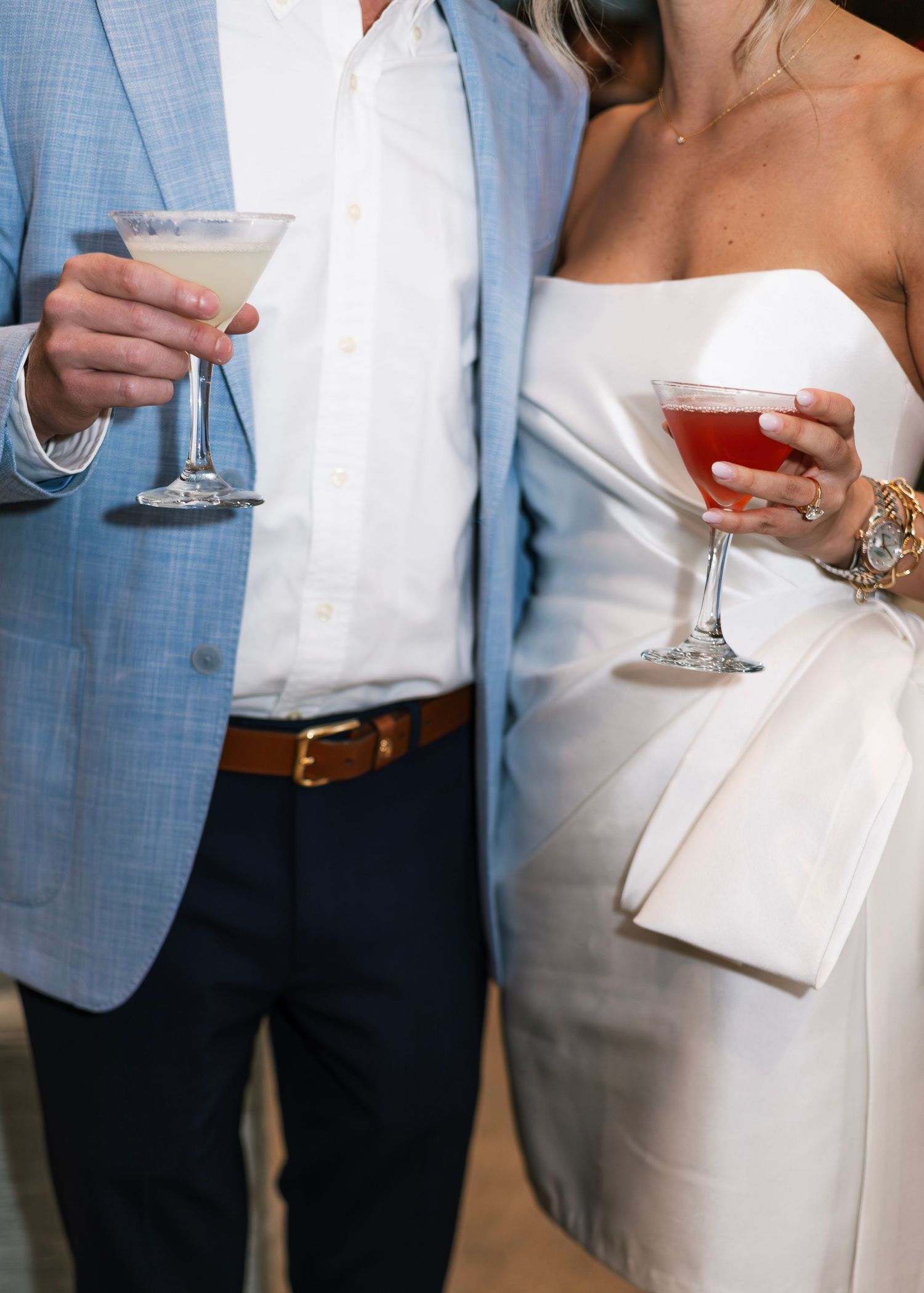 A couple in formal attire holds wine glasses at an elegant event, with the man wearing a light blue blazer.