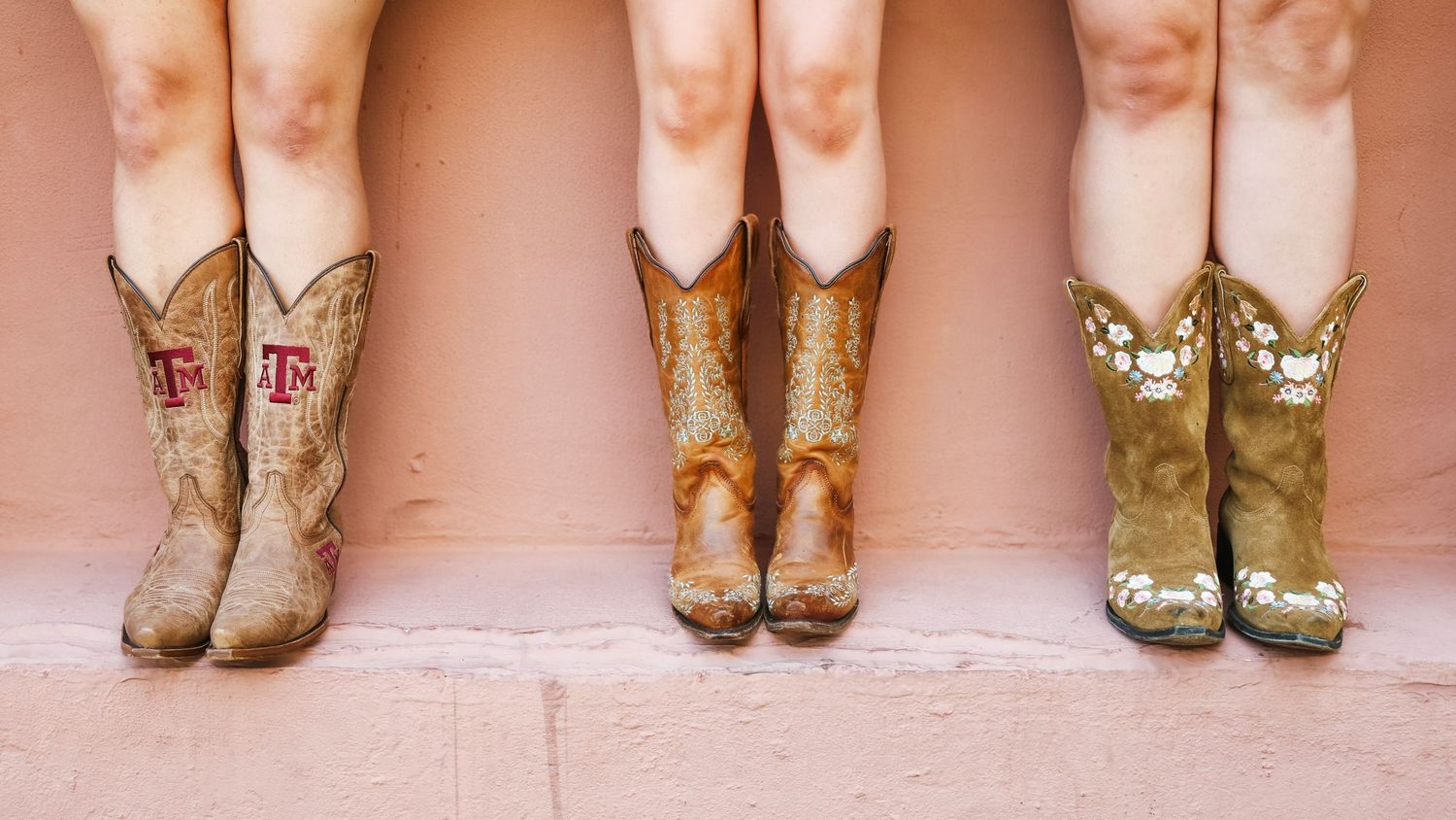 Row of cowboy boots lined up against a pink wall showing different styles and designs.
