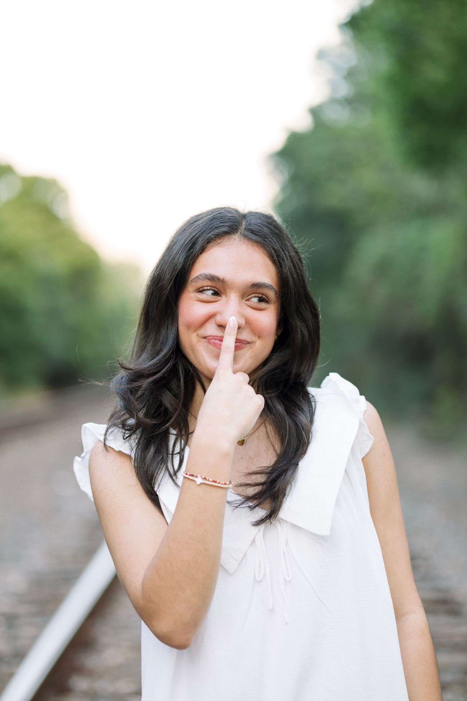Young woman in white dress posing playfully on railroad tracks, finger on nose, smiling outdoors.