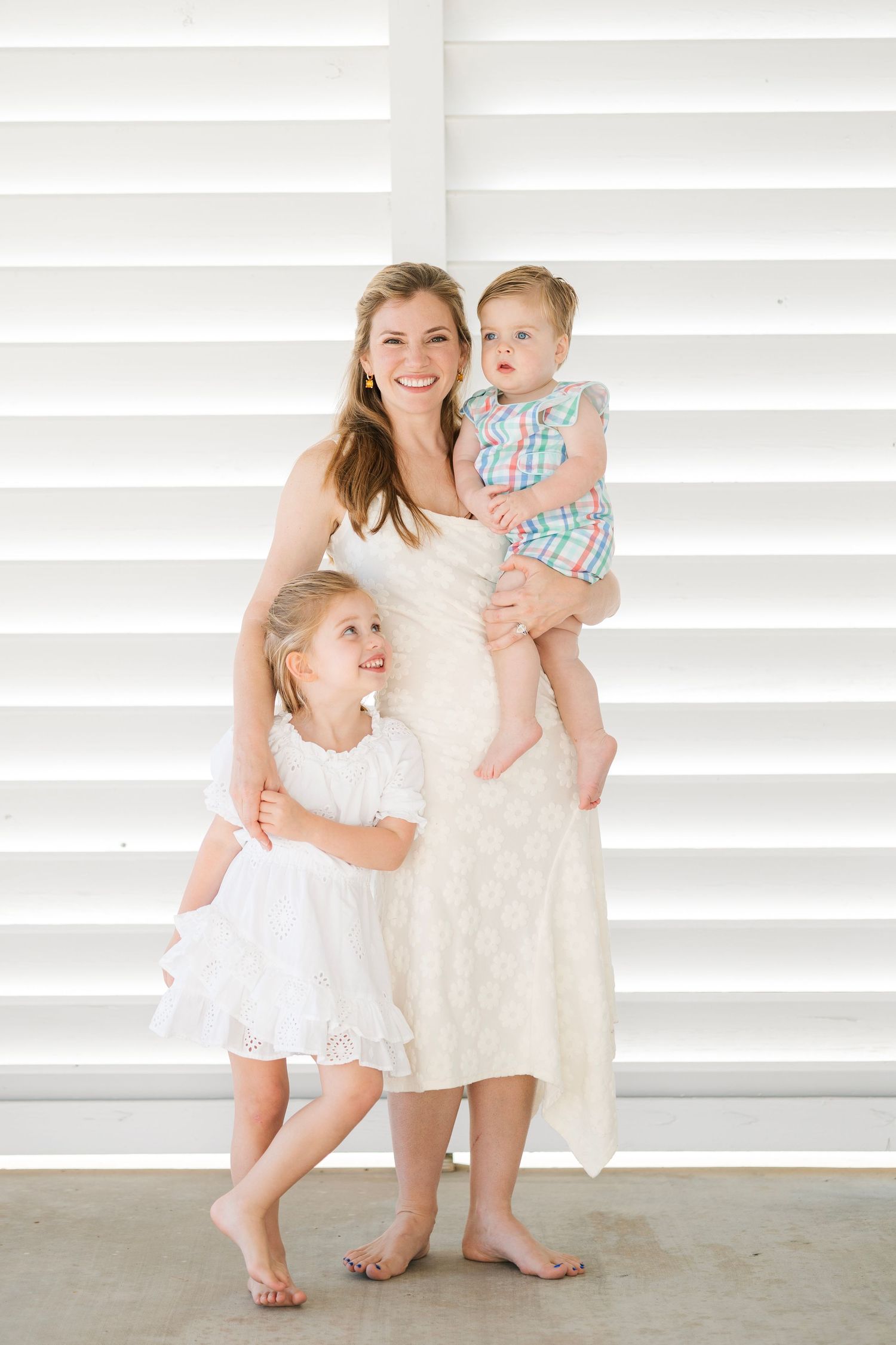 A smiling mother in a white dress holds her toddler while her young daughter hugs her leg indoors.