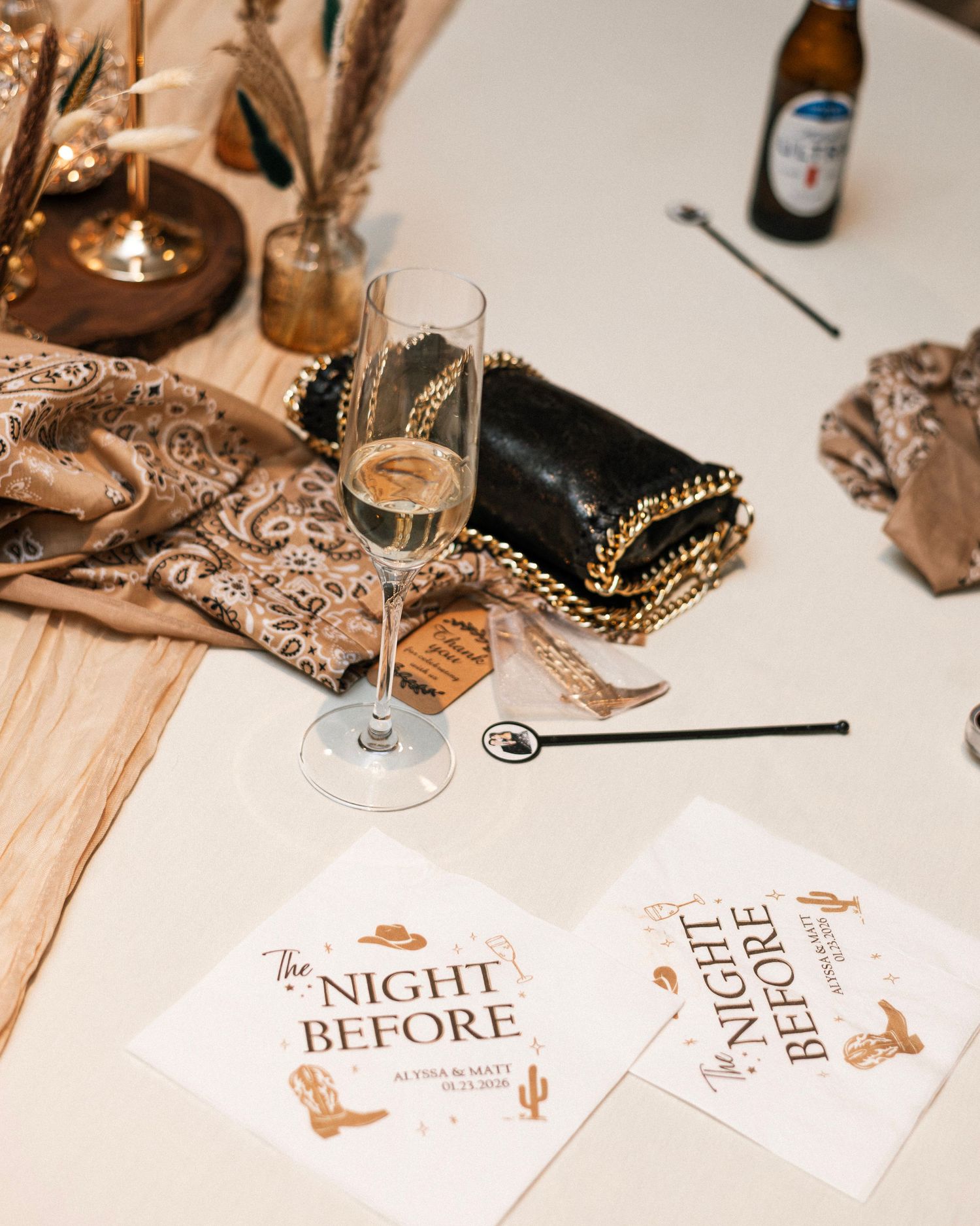 Elegant bachelorette party table with champagne flute, cowboy-themed napkins reading The Night Before, beer bottle, and paisley scarf.