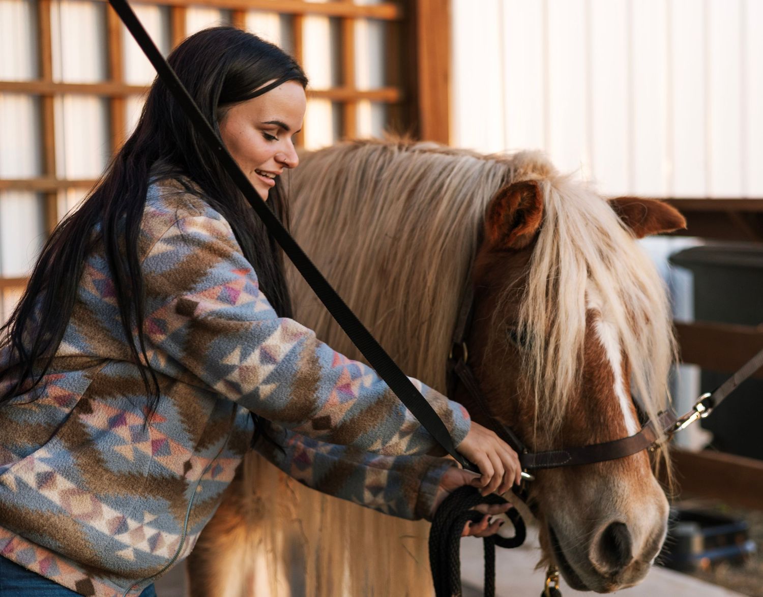 Woman in patterned jacket smiling while grooming a fluffy mane horse in a stable.