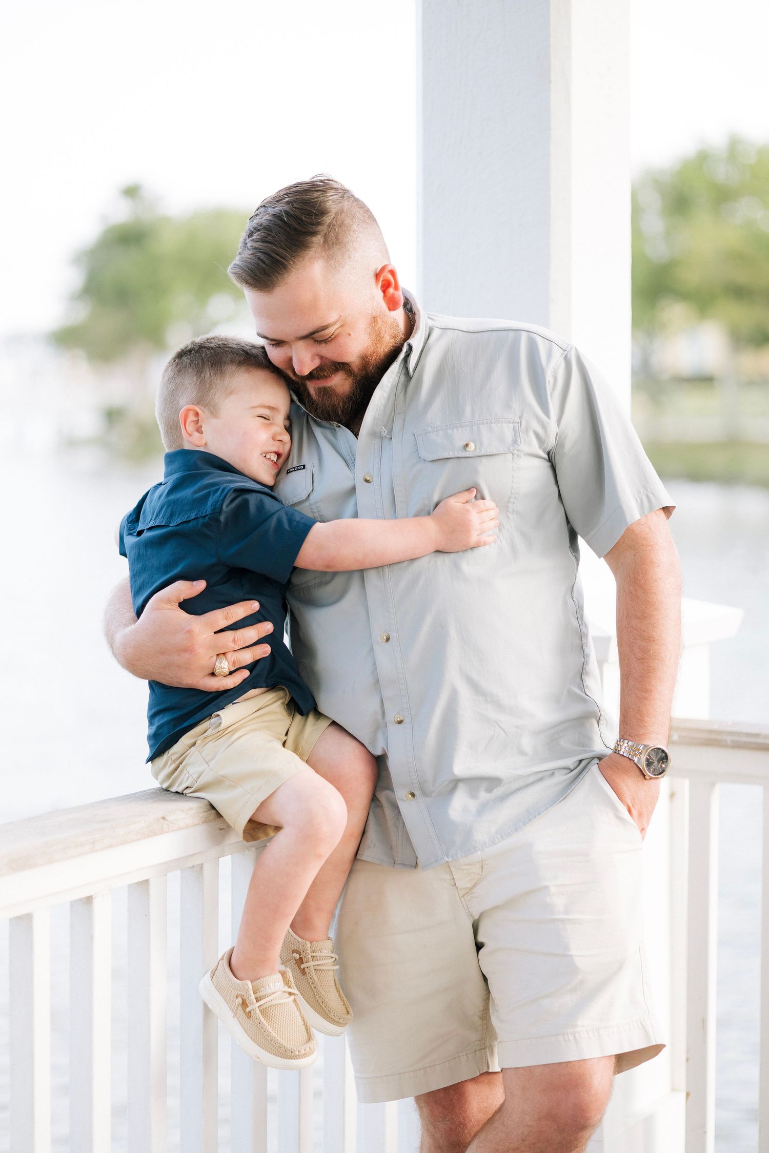 A father and young son share a tender hug on a waterfront porch, both smiling in casual summer outfits.